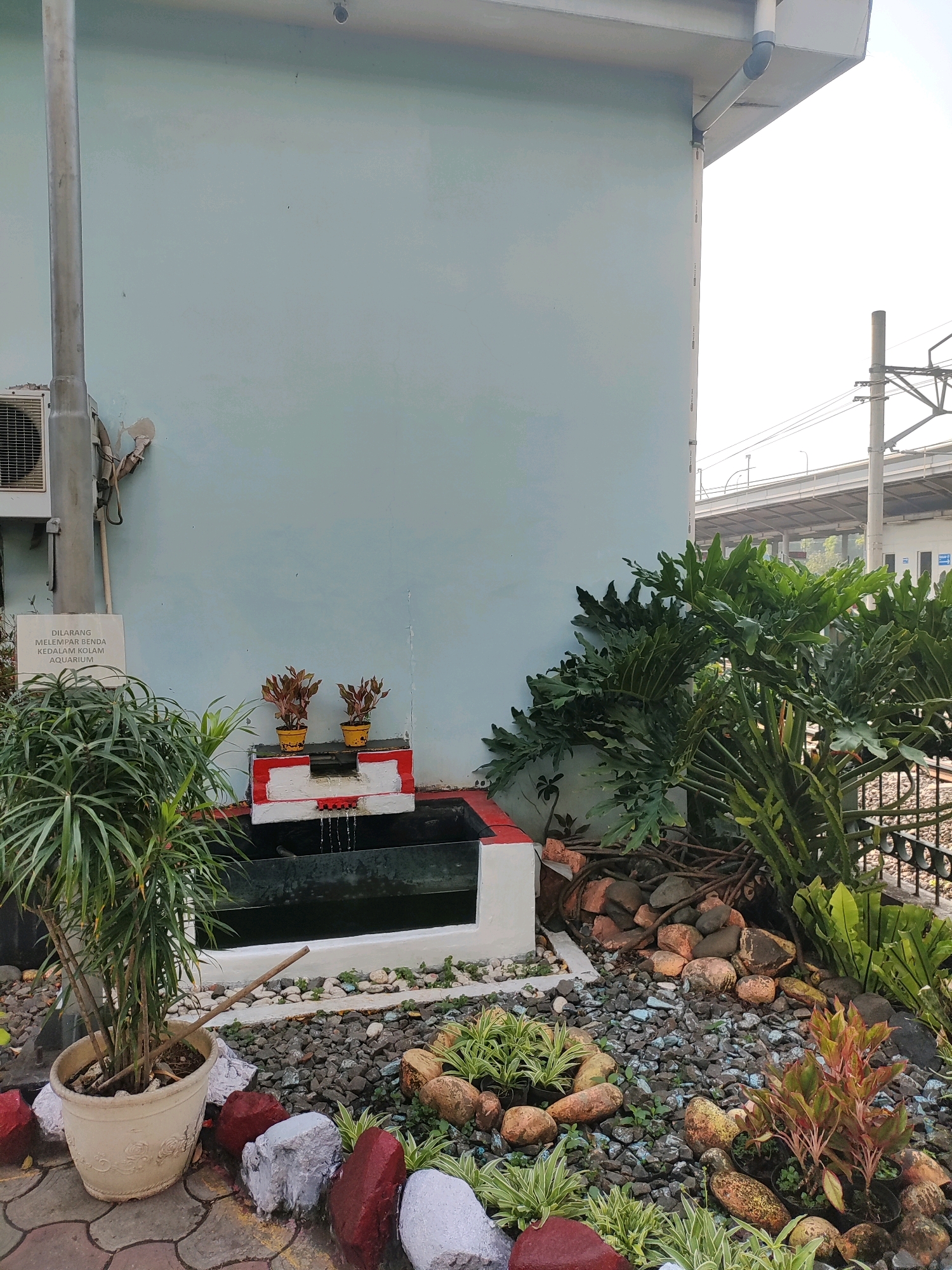 An outdoor, ground-level, slightly high-angle shot captures a small, ornamental water feature with cascading water into a dark pool. The water feature is adorned with a red and white border and two yellow potted plants. Surrounding the water feature is a garden bed filled with dark gray gravel, various sized rocks, and small green and reddish plants. To the left, a large potted plant with thin, green leaves stands beside a light blue wall. The wall has a sign with Indonesian text and an air conditioning unit mounted to it. To the right of the water feature, a large plant with broad, green, leafy fronds extends towards the foreground. In the background, a railway track and an elevated structure with power lines are visible against a bright sky. The overall scene depicts a small, decorative garden area next to a building.