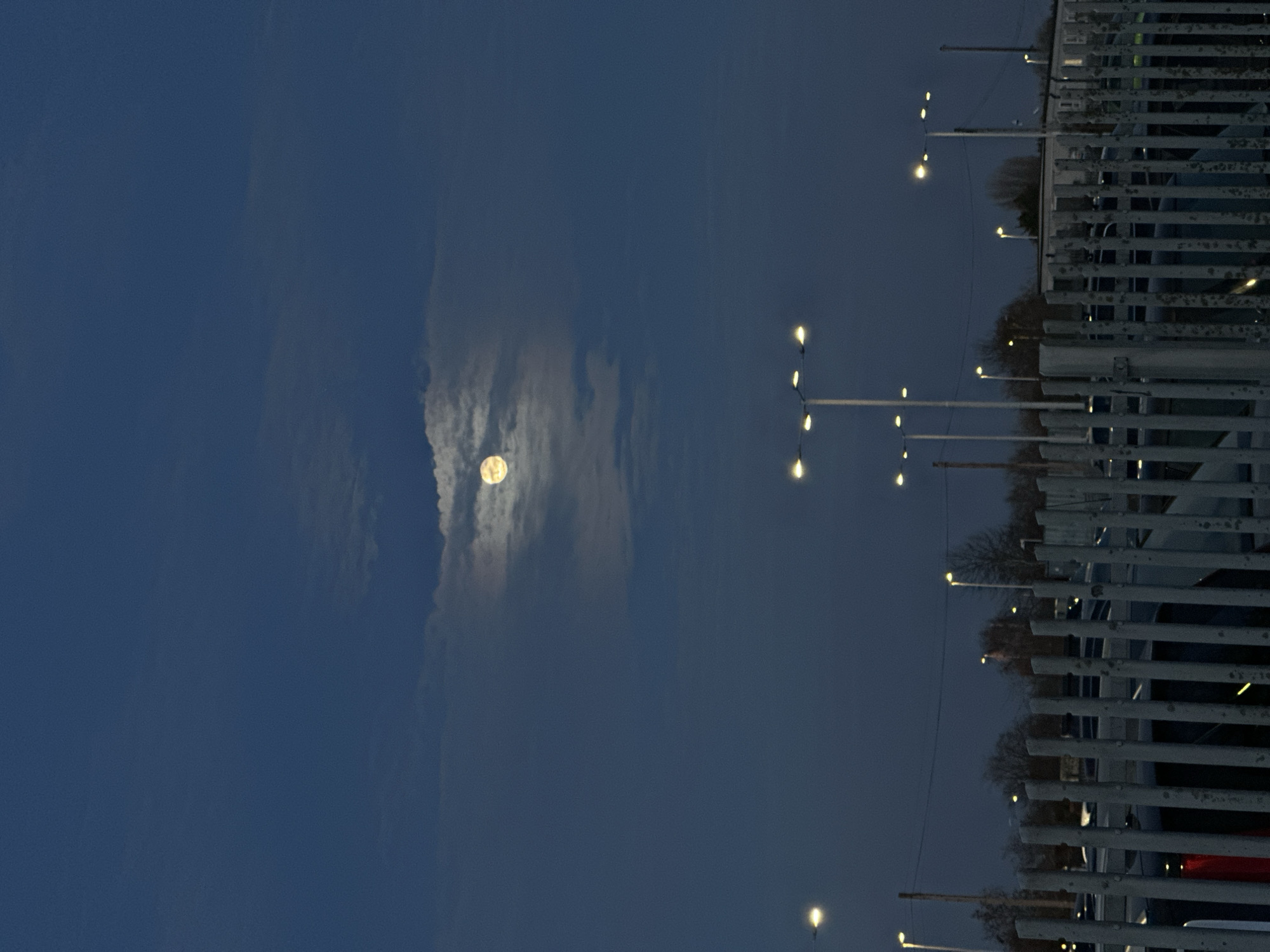 The image displays an outdoor scene at dusk or night in Sevenoaks, United Kingdom. A prominent, nearly full, yellow-white moon is visible centrally in the dark blue sky, partially obscured by thin, white clouds. Multiple streetlights with illuminated yellow-white bulbs are dispersed across the midground and background. The lower portion of the frame is dominated by a light grey, vertical palisade-style metal fence. Behind the fence, dark-colored vehicles are visible in what appears to be a parking area or car lot. Bare deciduous trees and bushes are present in the background, primarily on the left and center-right. A low, light-colored building with dark windows is partially discernible in the far background on the right side. No individuals are present in the scene.
