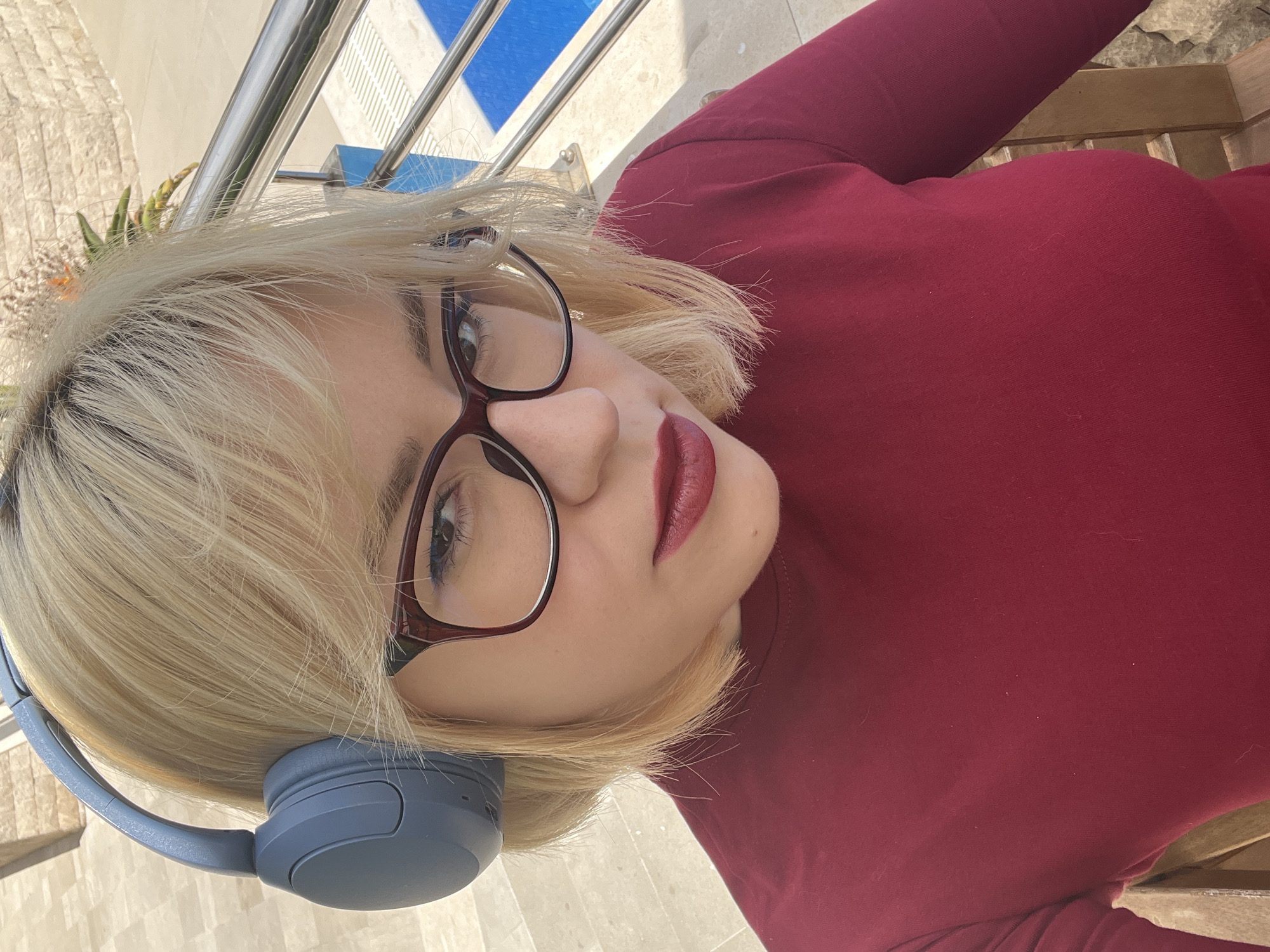 A woman with blonde hair, wearing glasses and headphones, is posing for a photo outdoors. She is wearing a maroon top. The background includes a blue and white tiled area with a metal railing. The photo appears to be taken in Boreti, Montenegro.