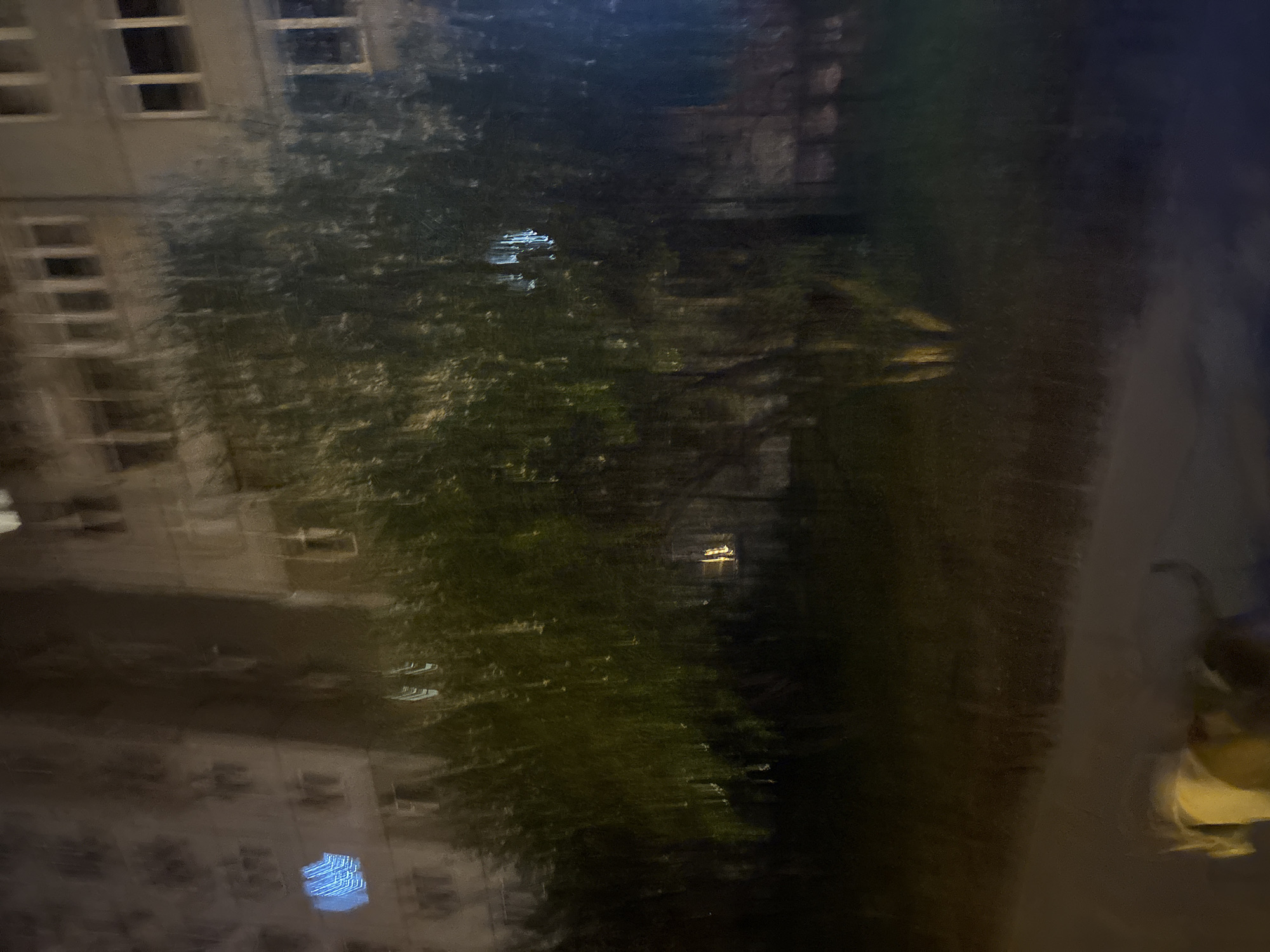 This blurry, low-light image, taken in Krasnoyarsk, Russia, appears to be a photograph captured through a window, likely at night or dusk, with significant motion blur or camera shake.
On the left side of the frame, a light-colored building with multiple dark, rectangular windows is visible, suggesting it's unlit from within or the exterior is dimly lit. A large, dense area of dark green foliage, possibly a tree or bushes, occupies the central and upper-right portions of the image.
Several bright, artificial light sources or reflections are scattered throughout the scene. A prominent warm-toned rectangular light is noticeable amidst the upper foliage, while a smaller, similar light appears further to the right. In the bottom-left area, a distinct blue rectangular light displaying a grid-like pattern is reflected on a surface, most likely the window pane through which the photo was taken, overlapping with the reflected image of the building. This reflection indicates an indoor light source.
The right side of the image is darker and less distinct, showing vague shapes that could be a path or ground. A highly blurred, yellowish shape on the far right edge suggests a distant, indistinct object or possibly a person. No specific activity is discernible, and no visible text is present.
