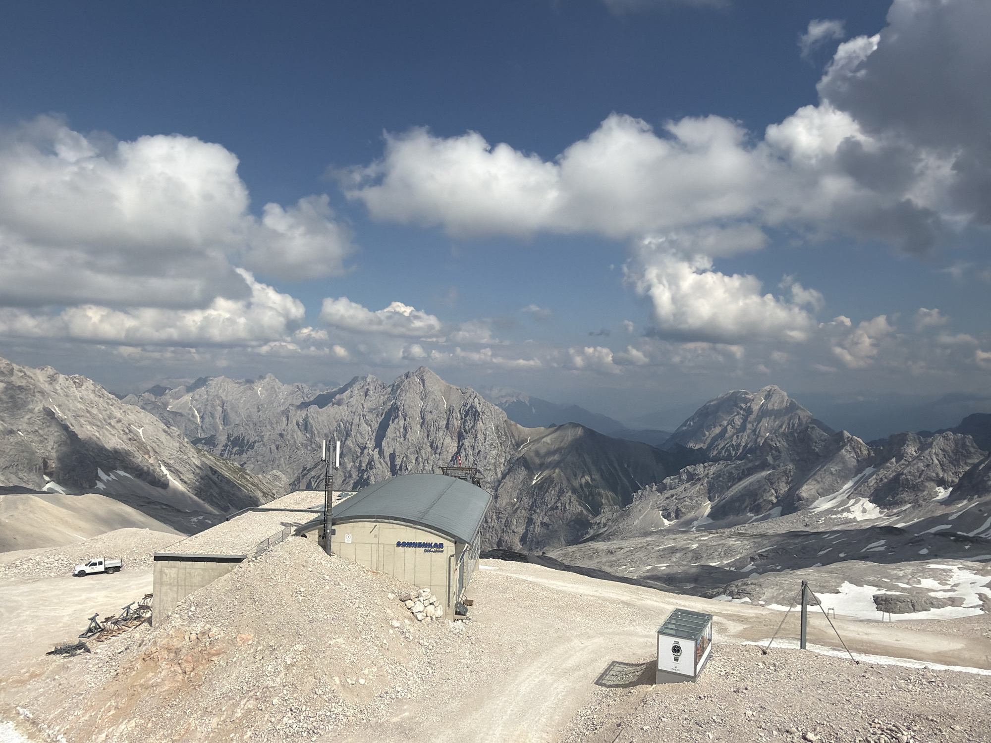 This media file captures a sweeping mountainous landscape under a dynamic sky. The scene is set in a high-altitude, rocky terrain, likely a mountain resort or observation point, given the presence of buildings and infrastructure. In the foreground, a gravelly path winds through the rocky ground.
Several structures are visible. A prominent building with a gray, curved roof and concrete walls stands near the center, featuring what appears to be a telecommunications tower on its roof. Text on this building reads "SONNENKAR" and "800-1600m". To the left of this building, a white pickup truck is parked on the path. In the lower right corner, a small, glass-enclosed display case is visible, with a watch face and the word "ROLEX" printed on it. Another similar structure is partially visible on the far right.
The background is dominated by majestic, rugged mountain ranges, their peaks sharp and imposing, with patches of snow still clinging to their slopes. The sky above is a vibrant blue, punctuated by a dramatic arrangement of white and gray cumulus clouds, suggesting a bright, possibly windy, day. The lighting indicates it is daytime, likely late morning or early afternoon, with shadows cast by the sun.
The overall impression is one of grandeur and isolation, highlighting the natural beauty of the alpine environment and the human structures that have been integrated into it. There are no people visible in the media file.