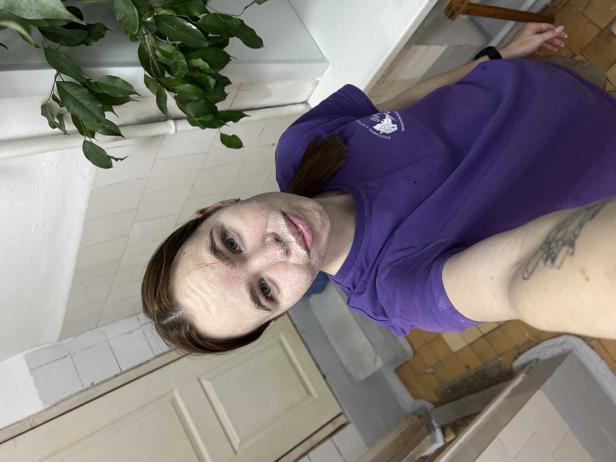 A woman with light skin and brown hair takes a selfie in what appears to be a bathroom or similar room with tiled walls and floors. She is wearing a purple T-shirt. Her face has a white substance, possibly a face mask. A green leafy plant is visible in the upper left corner. The tiles suggest a location within the city of Berezniki, Russia.