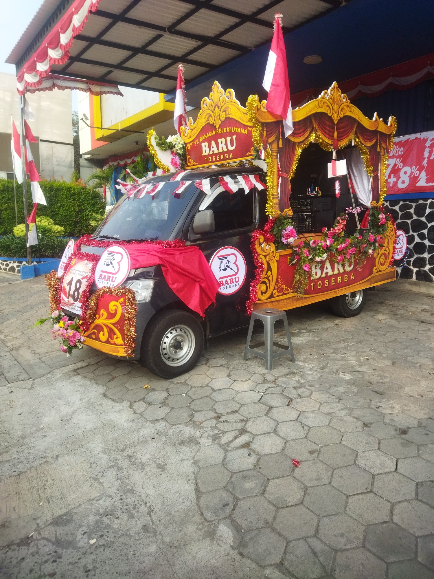 A decorated black minivan, possibly a Wuling Confero, is parked outdoors on a hexagonal paver surface. The vehicle is adorned with red and white Indonesian flags, red fabric, and floral arrangements. A golden archway with red curtains and decorative tassels is mounted on the rear of the vehicle, featuring the text "PT. BAHAGIA REJEKI UTAMA BARU TOSERBA" and additional floral decorations. A circular decal with a stylized "B" inside a circle and the text "BARU TOSERBA" is visible on the side of the van, along with a larger circular decal displaying the number "18" and similar branding. A small gray plastic stool is placed to the right of the front of the van. In the background, a building with yellow and blue accents and a covered walkway with red and white valances are visible. A black stone wall with a red banner displaying "Disc. 7% +8%" is on the far right. The scene suggests a festive event or parade, likely in Wonogiri, Indonesia, given the presence of Indonesian flags and the branding on the vehicle.
