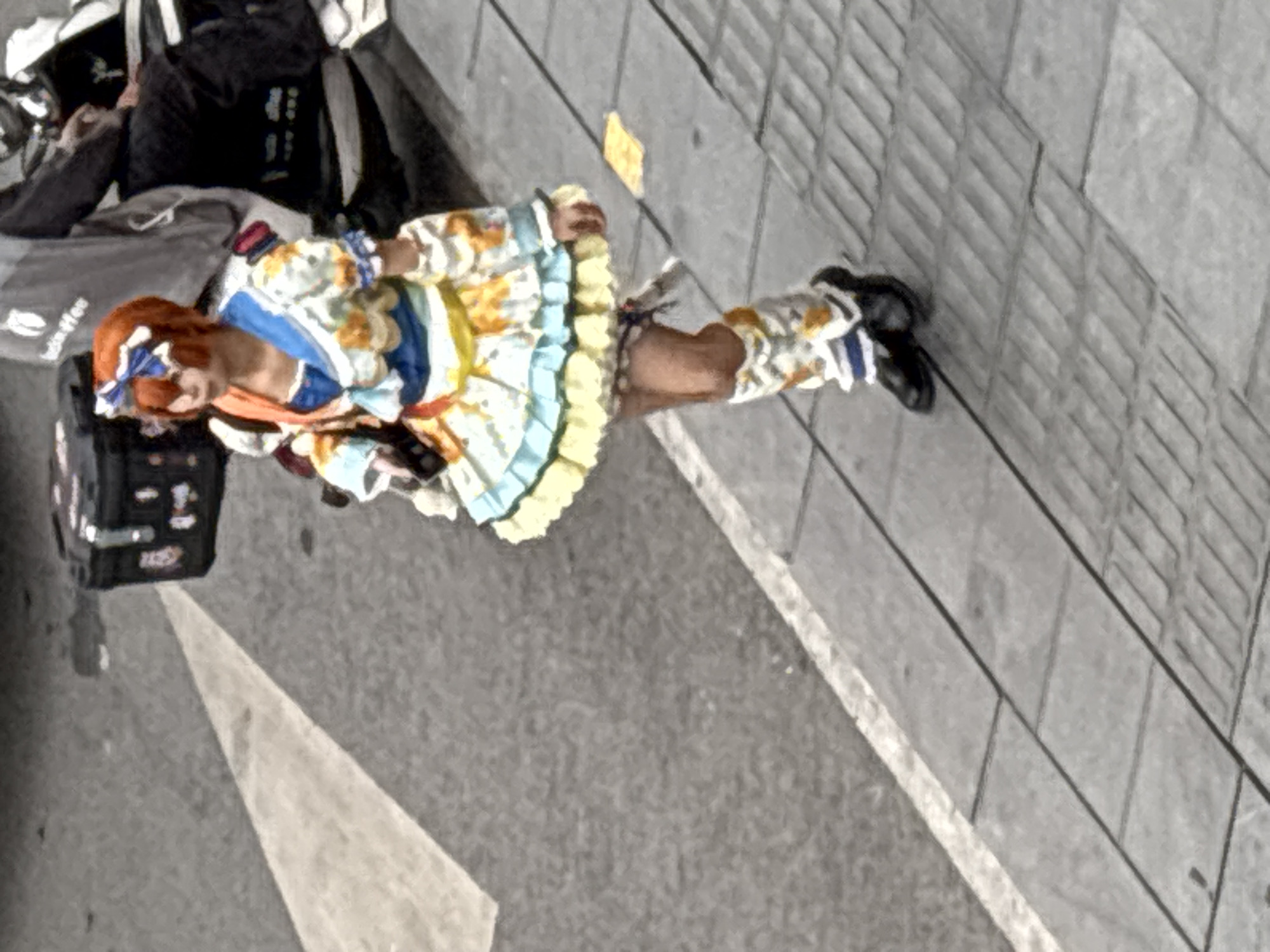 A person in cosplay is walking on a sidewalk in Jiulongpo District, China. The cosplayer wears a colorful dress, orange wig, and black shoes. A delivery bag and backpack are on the ground next to the cosplayer. The sidewalk is grey and made of rectangular slabs. A white line runs along the edge of the sidewalk, next to a grey road. A yellow rectangular object is lying on the sidewalk.