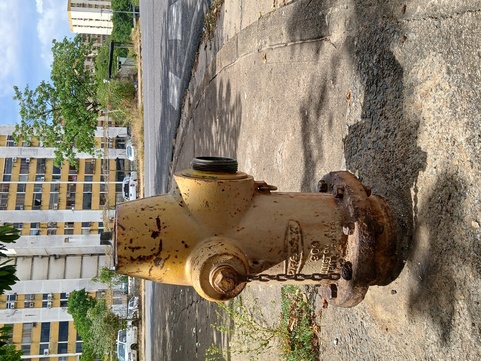 The image captures a yellow fire hydrant located on a concrete sidewalk in Ciudad Guayana, Venezuela. The hydrant shows signs of wear and rust, suggesting it is not new. A chain is attached to a cap or valve on the hydrant. The sidewalk abuts an asphalt street. In the background, several high-rise buildings are visible, typical of urban residential areas. One building has a distinctive yellow and white facade with numerous air conditioning units visible in the windows. Trees are interspersed between the buildings and the street. The image is taken during the daytime under natural light.