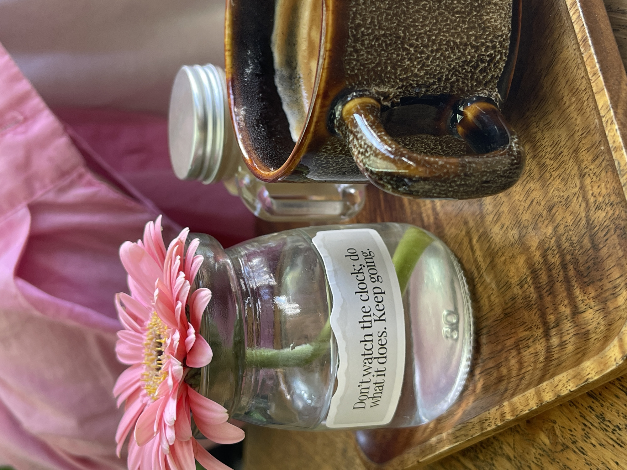 The image features a still life arrangement set on a wooden tray. A pink gerbera daisy is placed in a clear glass jar. The jar has a white label with the text "Don't watch the clock; do what it does. Keep going." To the upper right, a textured, brown ceramic mug rests partially on the tray and another small glass jar. A pink cloth provides a backdrop on the left side. The scene's specific location in Da Lat, Vietnam, cannot be definitively ascertained from the visual information provided.