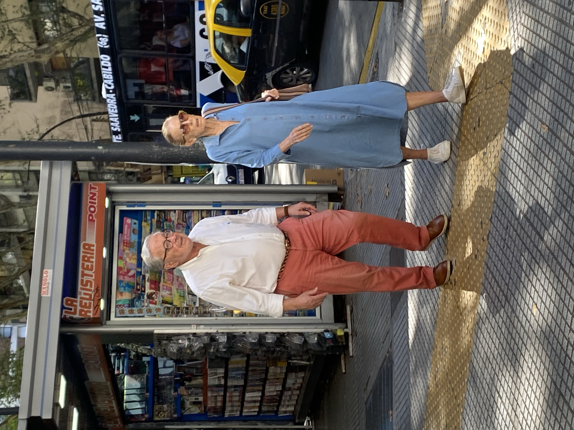 The image captures a scene on the streets of Buenos Aires, Argentina. An elderly couple is seen on the sidewalk. The woman, wearing a blue dress, sunglasses, and white sneakers, walks past a man wearing a white shirt, red pants, and brown leather shoes. Behind them, a bus and a taxi are parked. To their left, there is a storefront named "LA REGISTERIA," which appears to be a kiosk selling newspapers and magazines. The signage on the bus includes "TE. SAAVEDRA CABILDO (68) AV. SA," indicating the bus route or location.