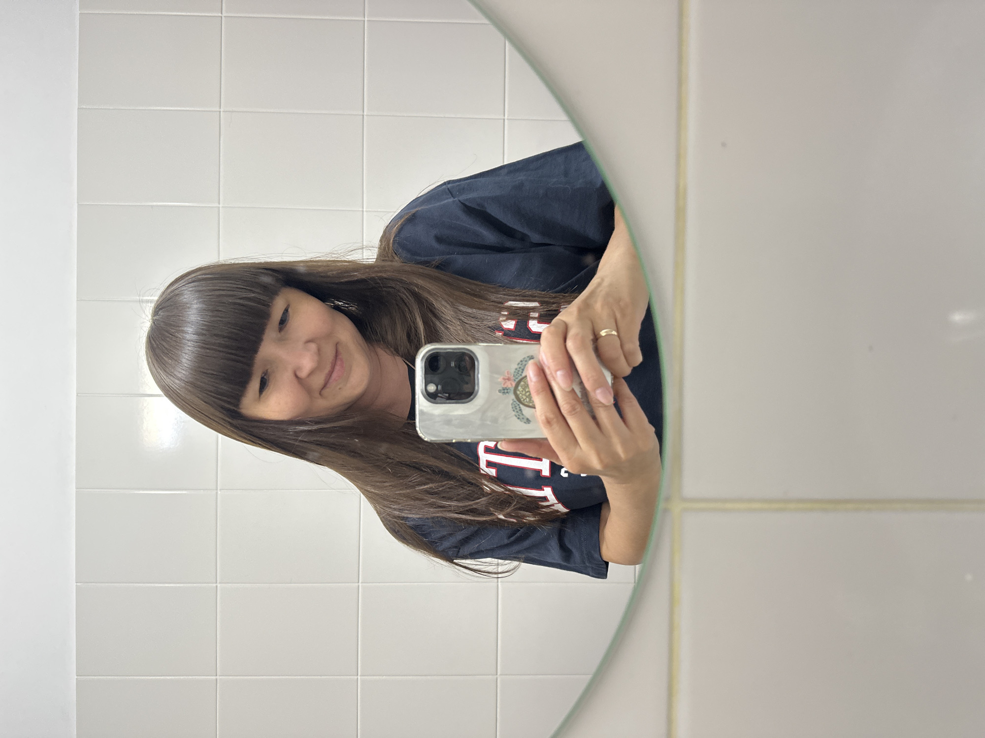 A woman with shoulder-length brown hair and bangs is taking a selfie in a bathroom mirror. She is wearing a dark blue t-shirt with white and red lettering. She is holding a smartphone in her right hand, which has a clear case with a design of a turtle and flowers. A gold ring is visible on her left ring finger. The background of the mirror is white tile. The lower part of the image shows the reflection of the woman from the waist down, and the bottom of the mirror frame. The scene is indoors, likely in a residential bathroom. There is no indication of the city of Chaikovsky, Russia, being present in the image.
