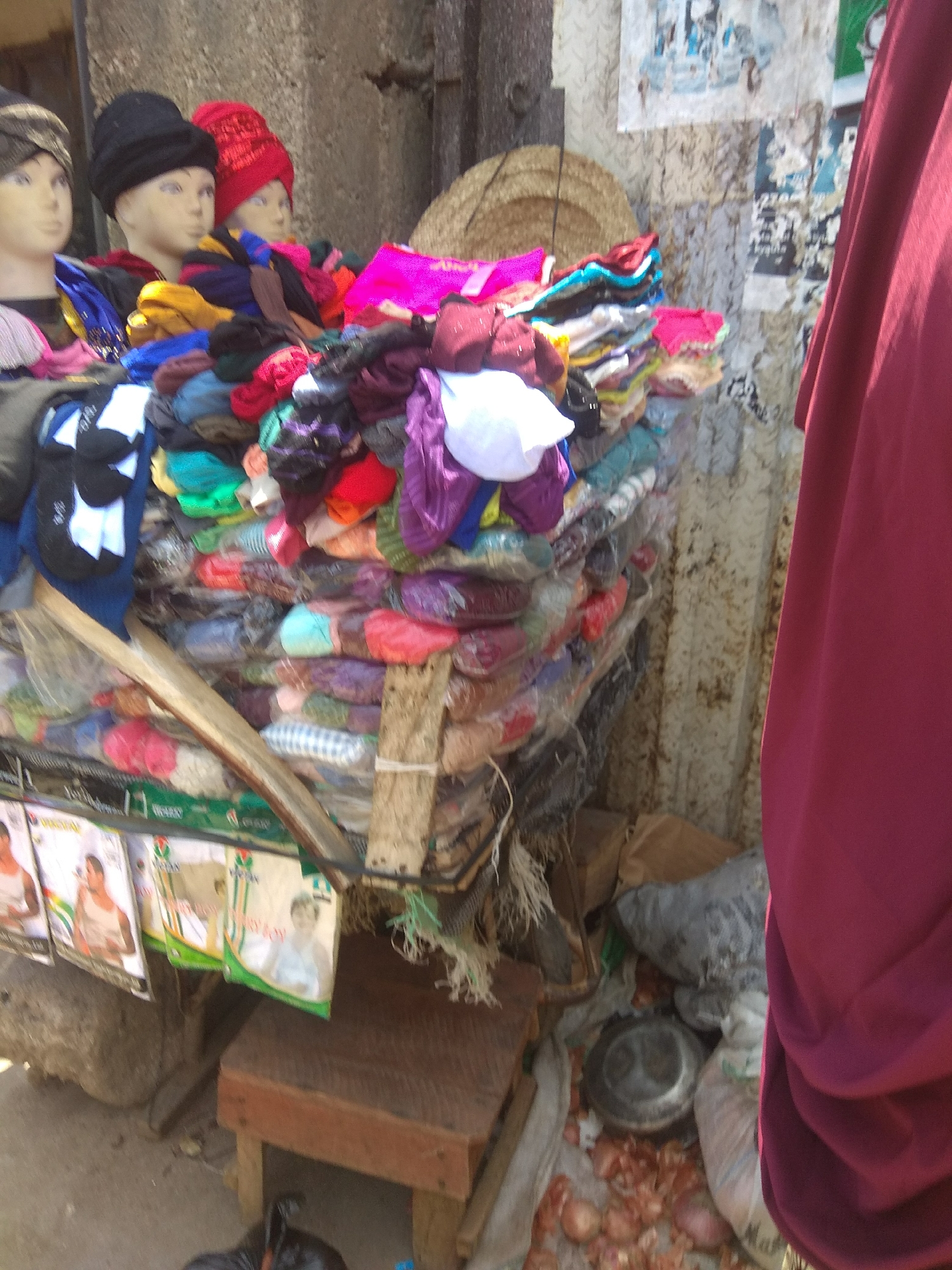 The image depicts an outdoor market stall in Jalingo, Nigeria, likely during the daytime. The stall is laden with a colorful assortment of knitted hats and other clothing items, predominantly piled high on wooden shelves. In the foreground, three mannequins are visible, two with dark headwear and one with a red knitted hat. The background features a textured, light-colored wall with some posters or advertisements pasted on it. To the right, a person is partially visible, draped in a dark red fabric, possibly a garment or a curtain. Below the stall, there are some bagged items and scattered onions. The overall scene suggests a bustling, everyday market environment with a focus on the sale of winter wear.