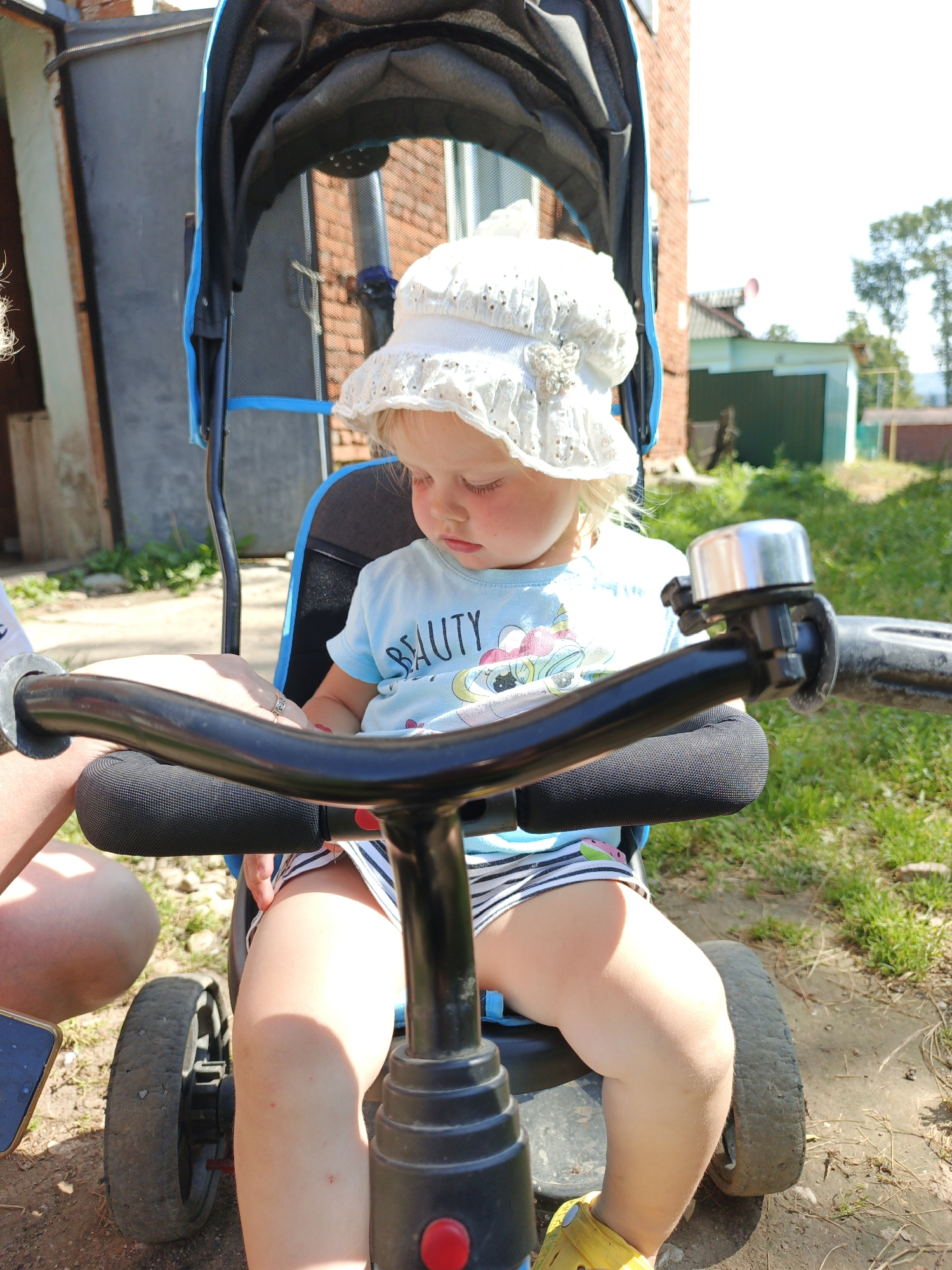 In Medvedyovka, Russia, a young Caucasian child with blonde hair is seated in a blue and black stroller. The child is wearing a white lace hat and a light blue t-shirt with a graphic print and the word "BEAUTY" on it, along with striped shorts. A hand, possibly belonging to an adult, is visible resting on the stroller's handlebar. A silver bicycle bell is attached to the handlebar. The stroller itself is a three-wheeled model with black grips and a black padded bar in front of the child. In the background, a brick building and a green fence are visible, along with trees and grass, suggesting an outdoor, possibly residential or rural, setting. The ground is a mix of dirt and grass.
