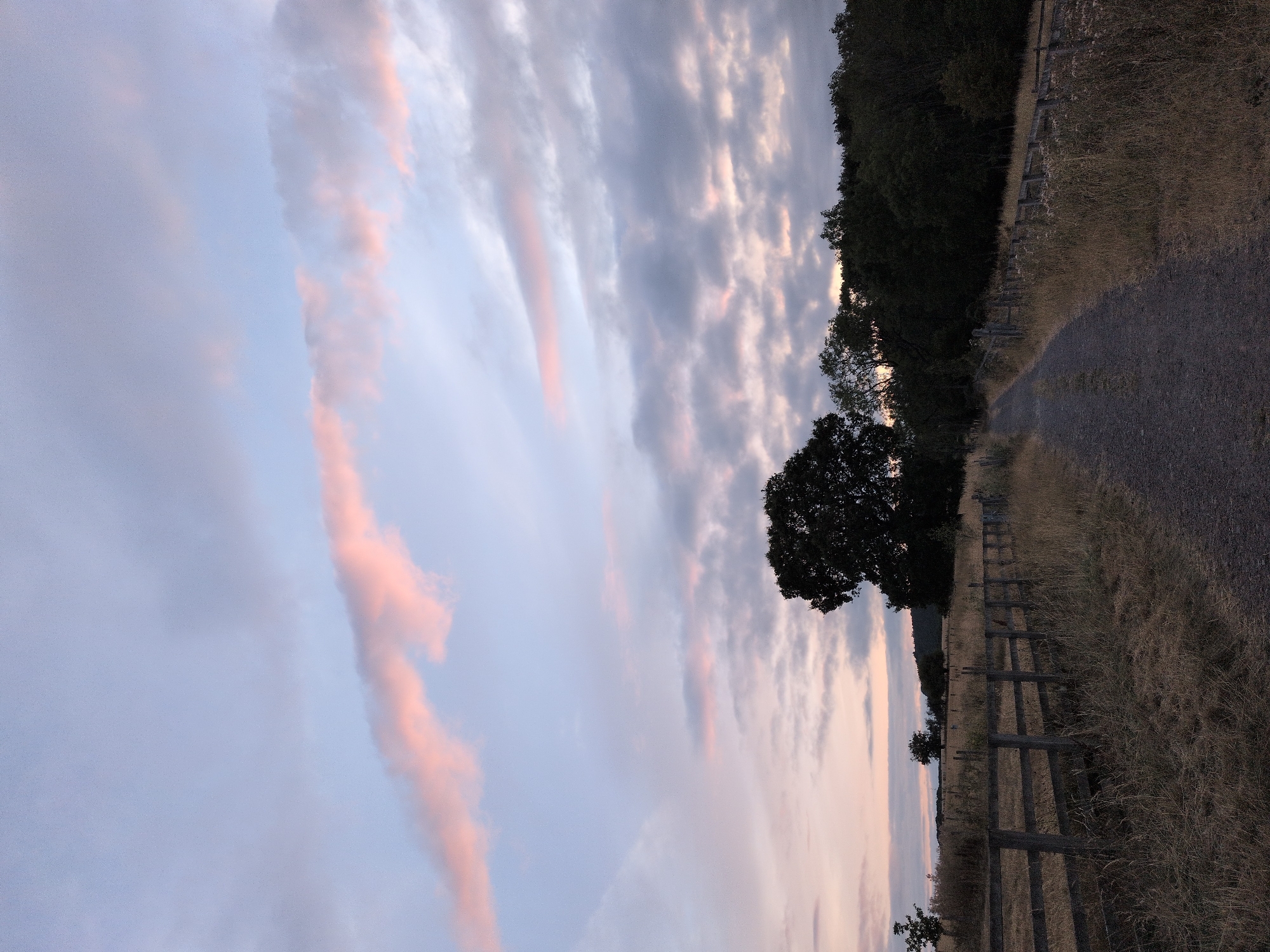 The image depicts a landscape scene at dusk in Little Wenlock, United Kingdom. In the foreground, a dirt path runs towards a dense line of trees and a rolling hill in the background. A wooden fence runs parallel to the path on the left side of the image, separating it from a field of dry grass. The sky is filled with a mixture of grey and pink clouds, suggesting the setting of the sun. The overall impression is one of a tranquil rural setting.