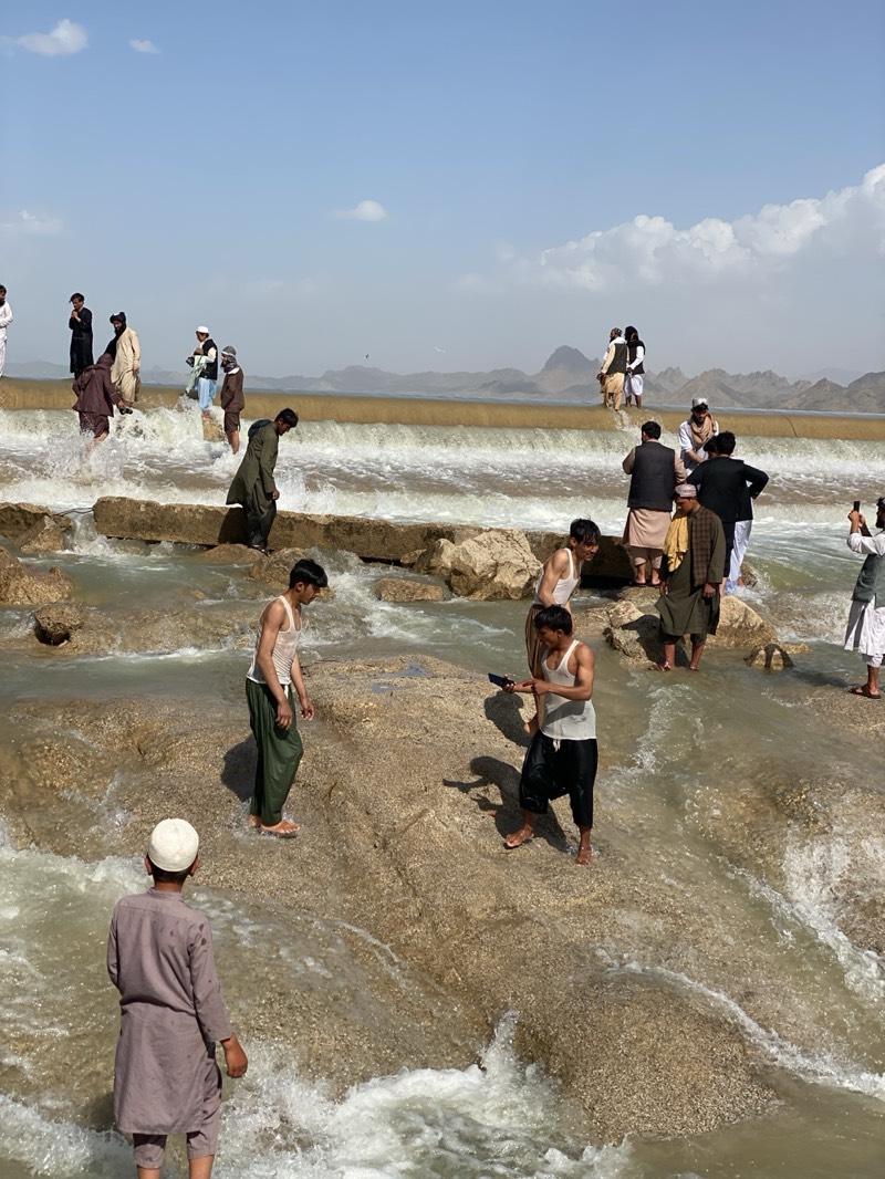 The image shows a group of people at Band-e Amir National Park, near the city of Kim, Afghanistan. Several men are standing on or near a low dam/waterfall structure, with water flowing over it. Some are fully clothed in traditional Afghan clothing, including long robes and headwear, while others are wearing tank tops and loose pants. One man is holding a phone, possibly taking pictures. The water is shallow in the foreground and people are wading through it. In the background, there are hills or mountains. The sky is partly cloudy.