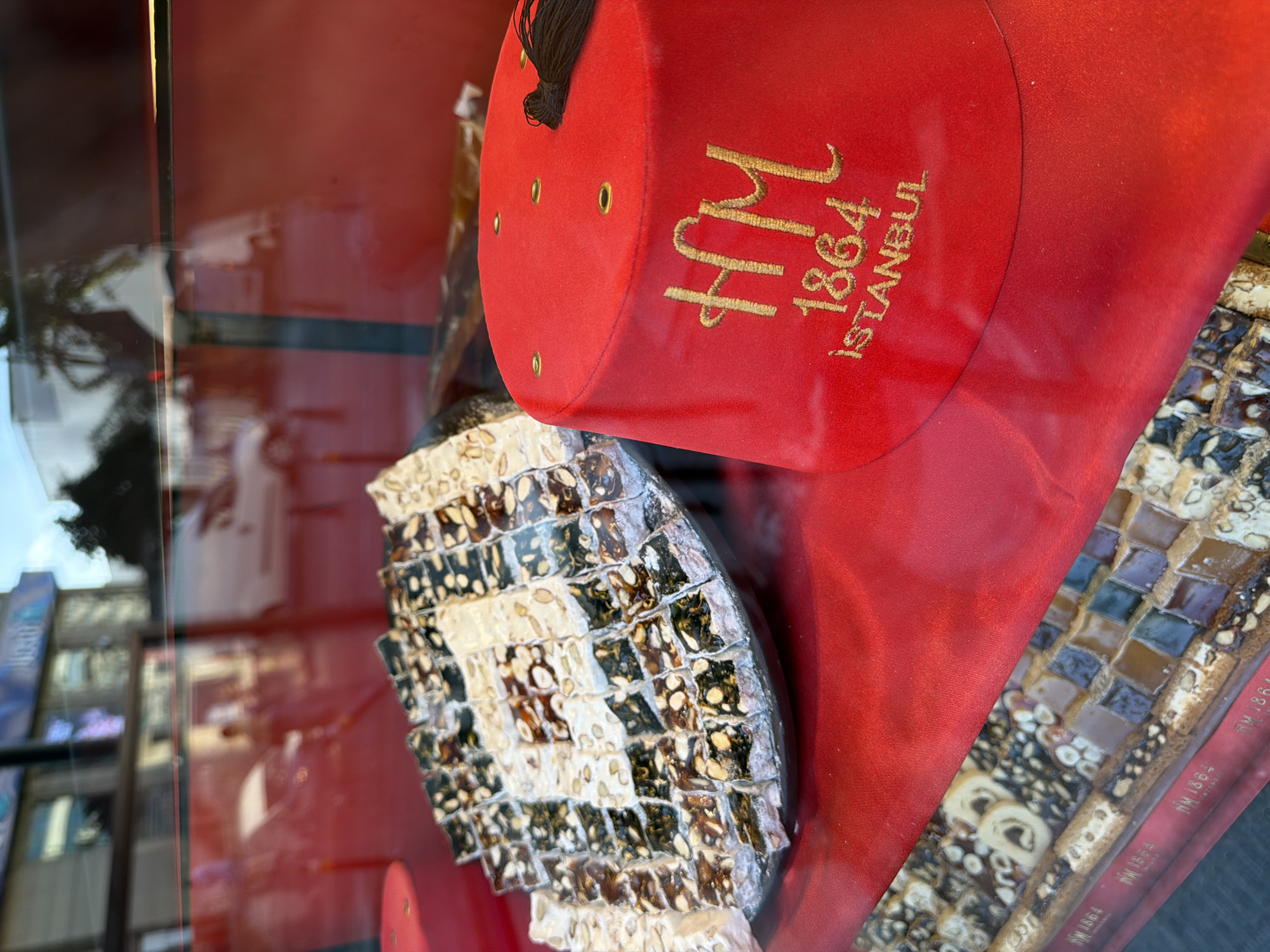 The image depicts a display window of a confectionery shop in Istanbul, Turkey. In the foreground, on the right, is a red fez hat with gold embroidery that reads "HM 1864 ISTANBUL". To the left of the hat, a large platter is filled with various types of Turkish delight, cut into small rectangular pieces. The Turkish delight has different colors and visible ingredients like nuts. Beneath this platter, another display case shows more rows of Turkish delight. The background shows reflections of a street scene, including vehicles and buildings, suggesting this is an outdoor shop display.