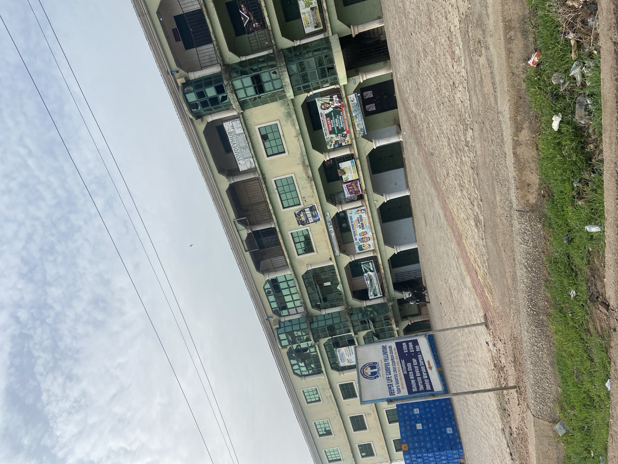 The image captures a multi-story building in Lokoja, Nigeria, seen from a slightly low angle, alongside a paved path and a grassy verge under an overcast sky. The building, painted in a light green or beige hue, features numerous square windows, some with a greenish tint, and several balconies. Some window panes appear to be broken or damaged. Various commercial and organizational banners and posters are affixed to the building's facade.
Prominently in the foreground, a blue and white signpost displays information for the "DEEPER LIFE CAMPUS FELLOWSHIP," indicating its connection to "Federal University Lokoja." The sign lists activities: "Monday Bible Study - 5:53pm," "Thursday Revival Hour - 5:50pm," and "Sunday Worship Service - 8am." Nearby, several blue crates are stacked on the ground.
Banners visible on the building advertise "YOUDEVHUB," "OMEGA FIRE," "FANCY PRESSURE NETWORK FOR GWWA," "USMAN ODODO," and "TEMI-T COUTURE," among other less decipherable text like "GREAT COMMUNITY ARABIA" and "ELEGANT GUY SUPERMART."
The paved pathway runs along the building, bordered by a strip of grass that contains scattered litter and debris. Overhead, multiple power lines traverse the light gray, cloudy sky. A small bird is faintly visible in the distance. The scene is static, depicting a daytime urban environment.