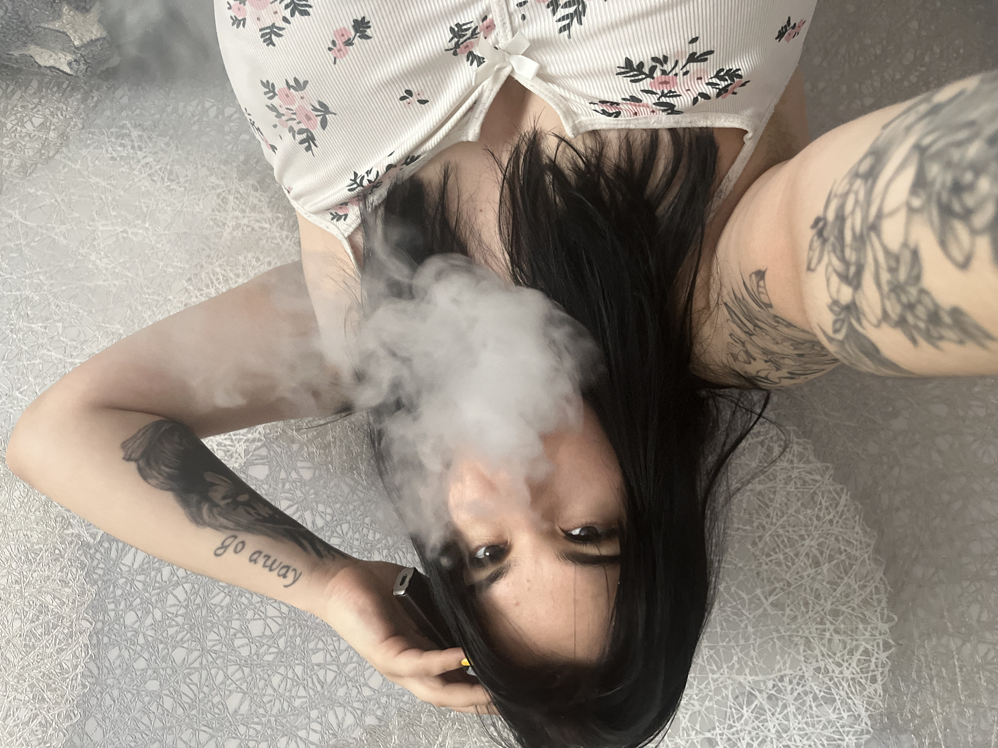 A woman is shown from a low angle, lying on her back on a surface with a white webbed pattern. She has dark hair, dark eyes, and is wearing a white top with a pink and black floral pattern. Her left arm is visible, showing a tattoo of a bird-like figure and the words "go away". Smoke is coming from her mouth. Her right arm is also tattooed with a design that covers most of her forearm. She is holding a vape device with her left hand. The location is unknown, and cannot be verified as being in Chaikovsky, Russia.