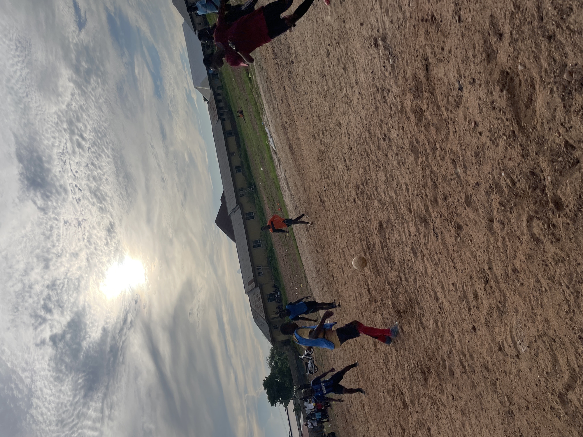 The image depicts an outdoor soccer game taking place on a dirt field in Lokoja, Nigeria. The sun is visible in the sky, partially obscured by clouds. Several young men are actively playing soccer, with one individual in a red shirt and black shorts running towards the right of the frame, and another in a blue and yellow jersey kicking a soccer ball on the left. Other players and spectators are present in the background, along with buildings that appear to be in a state of disrepair or under construction. A motorcycle is also parked on the left side of the image. The overall setting suggests a casual, informal sporting event in a developing urban or semi-urban environment.