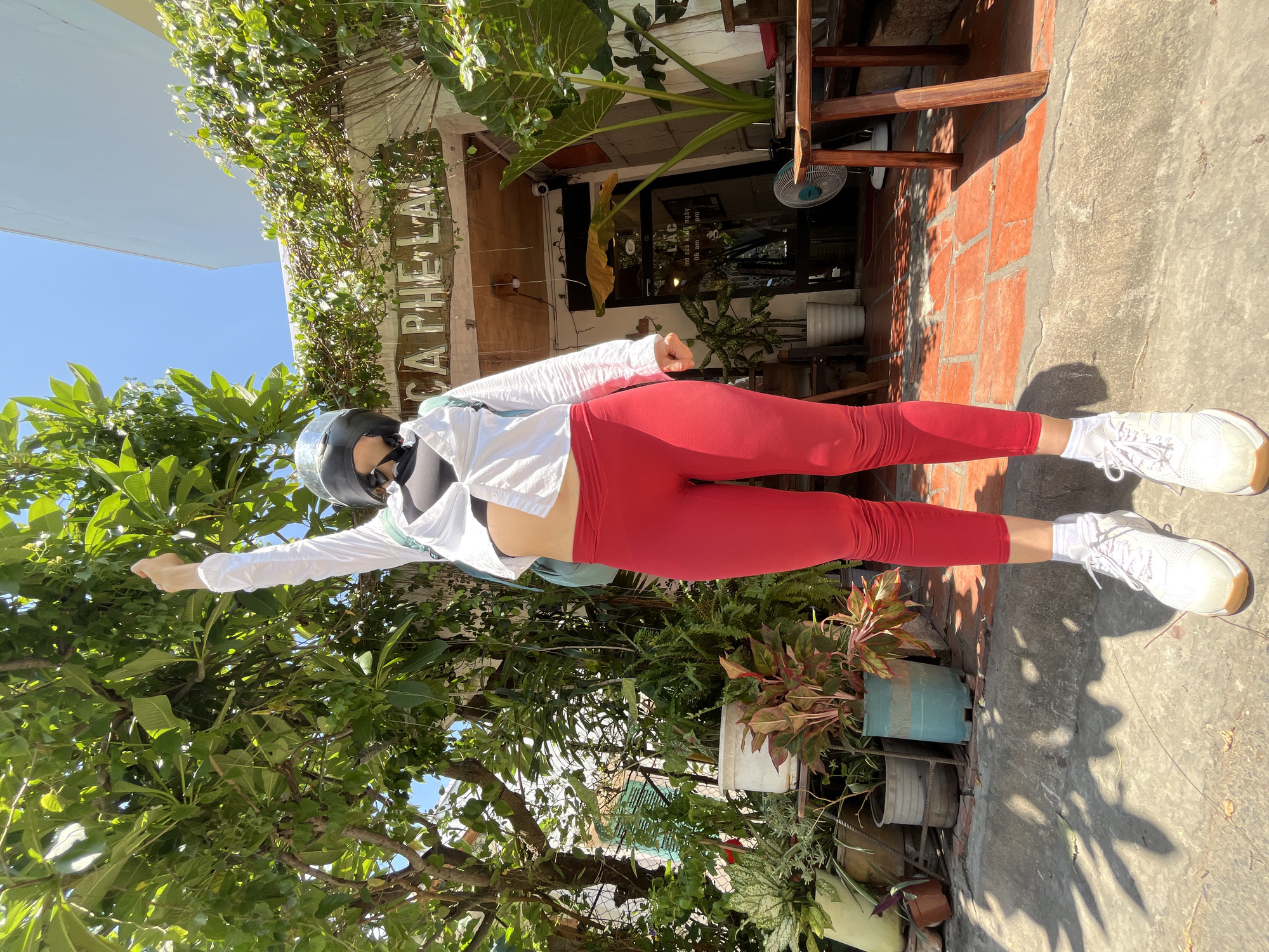 A person stands on an orange tiled pathway in front of a building with a sign reading "CA PHE LAN" in Phu Thuy Ward, Vietnam. The individual is wearing a black helmet, a white open-button shirt, a black sports bra, bright red leggings, and white athletic shoes with white socks. They are extending their right arm upwards, pointing towards the green foliage of a tree. The background features a building entrance with glass doors, plants in pots, outdoor seating, and a fan. The lighting suggests it is a sunny day with shadows cast on the ground.