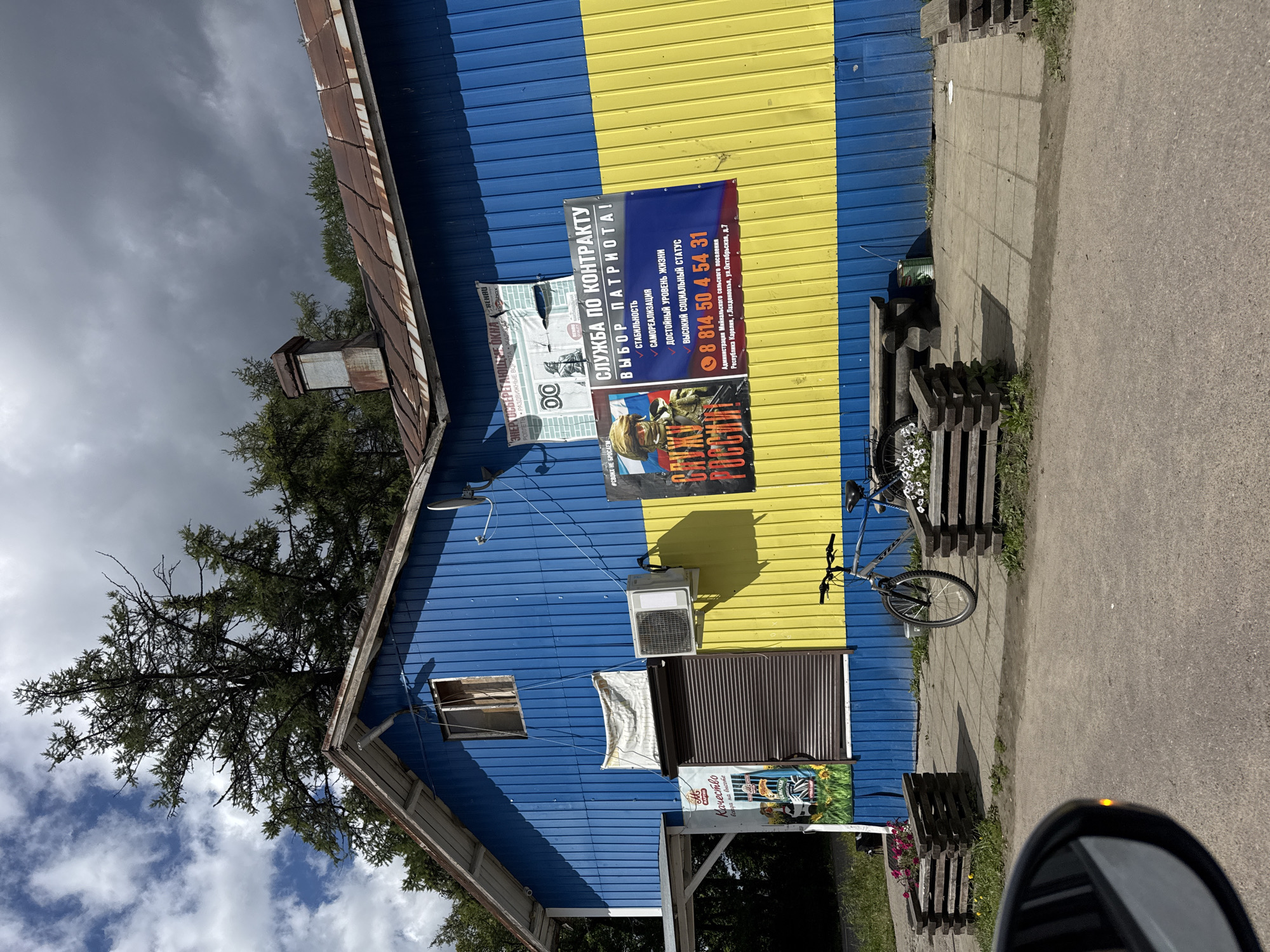 The image shows the exterior of a building painted in blue and yellow, evoking the Ukrainian flag. The building is a single-story structure with a rusty, corrugated metal roof and a prominent chimney. On the left side of the building, there is a covered walkway. Various posters are attached to the facade, including one advertising "SERVICE BY CONTRACT" and another with the text "SERVE RUSSIA!". A bicycle is parked in front of the building, next to wooden planters filled with flowers and a wooden bench. A satellite dish is mounted on the upper right side of the building. The environment appears to be outdoors, with trees and a cloudy sky visible in the background. The ground in front of the building is paved, and a car's side mirror is visible in the lower left corner of the frame, suggesting the photo was taken from a vehicle. The address "Administration of Mikhailovsky Municipal District, Republic of Karelia, Lakhdenpokhsky district, Oktyabrskaya str., 7" is visible on one of the posters. The presence of these posters and the address indicates the location is in the Mikhailovsky Municipal District of the Republic of Karelia, Russia, specifically on Oktyabrskaya Street, building 7, in the Lakhdenpokhsky district.