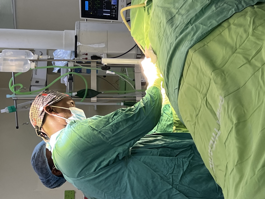 The scene is an operating room within a medical facility located in Nairobi, Kenya. Two medical professionals are present, engaged in a procedure. The foreground individual, presumed to be the primary surgeon, wears green surgical scrubs, a patterned surgical cap, a blue surgical mask, and yellow surgical gloves. This individual leans forward over a patient, whose body is primarily obscured by dark and light green surgical drapes, revealing a focused, brightly illuminated operative field. The surgeon's gloved hands are visible within this field, indicating active manipulation. A second individual, wearing blue surgical scrubs and a blue surgical cap, is partially visible behind and to the left of the primary surgeon, suggesting an assisting or observing role. In the background, an IV pole holds two intravenous fluid bags, while green medical tubing connects to what appears to be an anesthesia machine or ventilator with a dark green rebreather bag. A patient monitor displays vital signs, including a cardiac waveform and numerical data, on its screen. The environment is well-lit, with a specific, intense light source directed at the surgical area.