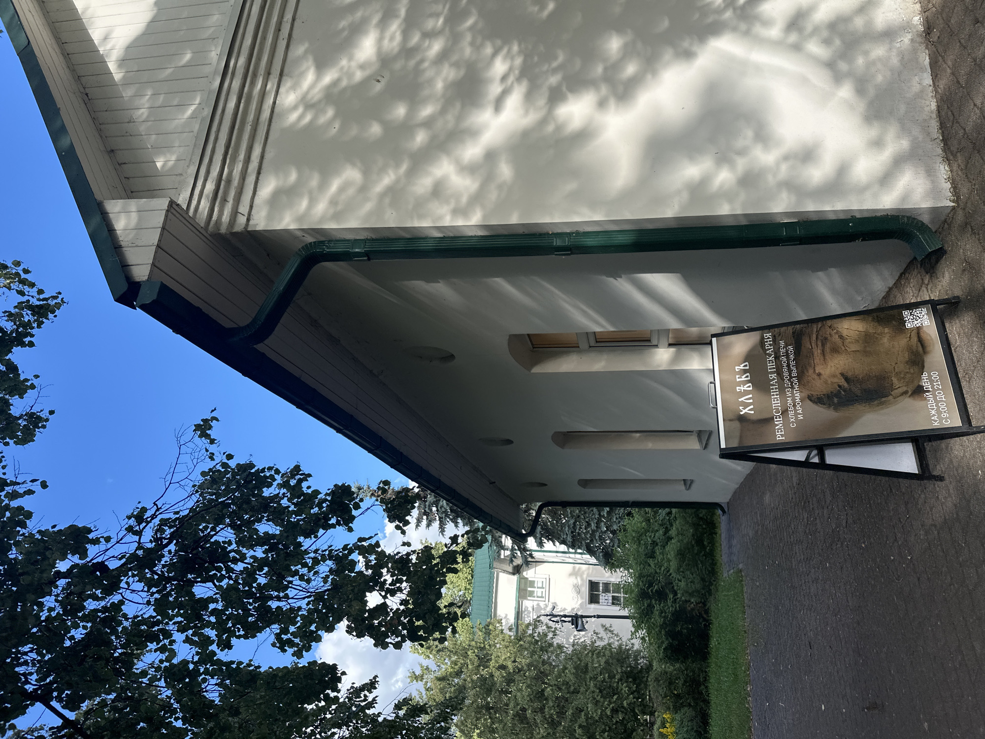 This is a photograph of a building with a green gutter and a white facade, dappled with shadows from trees. In the foreground, a black A-frame sign stands on a paved area. The sign displays Russian text and an image of bread. The text on the sign reads: "ХЛЕБЪ", "РЕМЕСЛЕННАЯ ПЕКАРНЯ", "С ХЛЕБОМ ИЗ ДРОВЯНОЙ ПЕЧИ И АРОМАТНОЙ ВЫПЕЧКОЙ", and "КАЖДЫЙ ДЕНЬ С 9:00 ДО 21:00". This translates to "BREAD", "ARTISAN BAKERY", "WITH BREAD FROM A WOOD-BURNING OVEN AND FRAGRANT PASTRIES", and "EVERY DAY FROM 9:00 TO 21:00". The sign indicates that this is an artisan bakery. Beyond the building, there are trees and another building visible. The sky is blue and clear, suggesting it is daytime with good weather. The scene appears to be outdoors, possibly in a town or city in Russia, given the location context. The overall impression is of a bright, sunny day outside a local establishment.