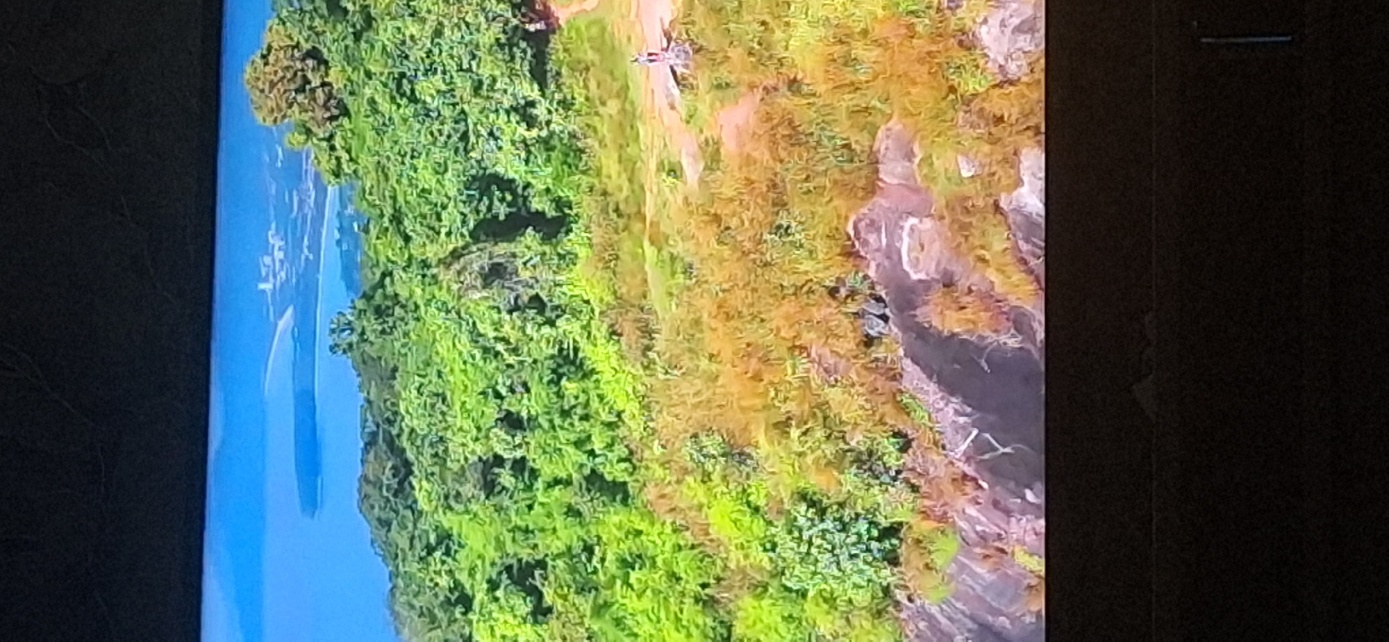 The image displays a panoramic view of a lush green hillside overlooking a body of water with a distant coastline featuring buildings. The hillside is covered with dense vegetation, transitioning from vibrant green foliage to dry, golden grasses towards the bottom. Rocky outcrops are visible on the slope. In the middle ground, on a dirt path, a person is standing near a stone structure. Further to the right, another person is standing on the path. The sky above is a bright blue. There are no discernible elements in this image that would place it within the city of Bila Tserkva, Ukraine.