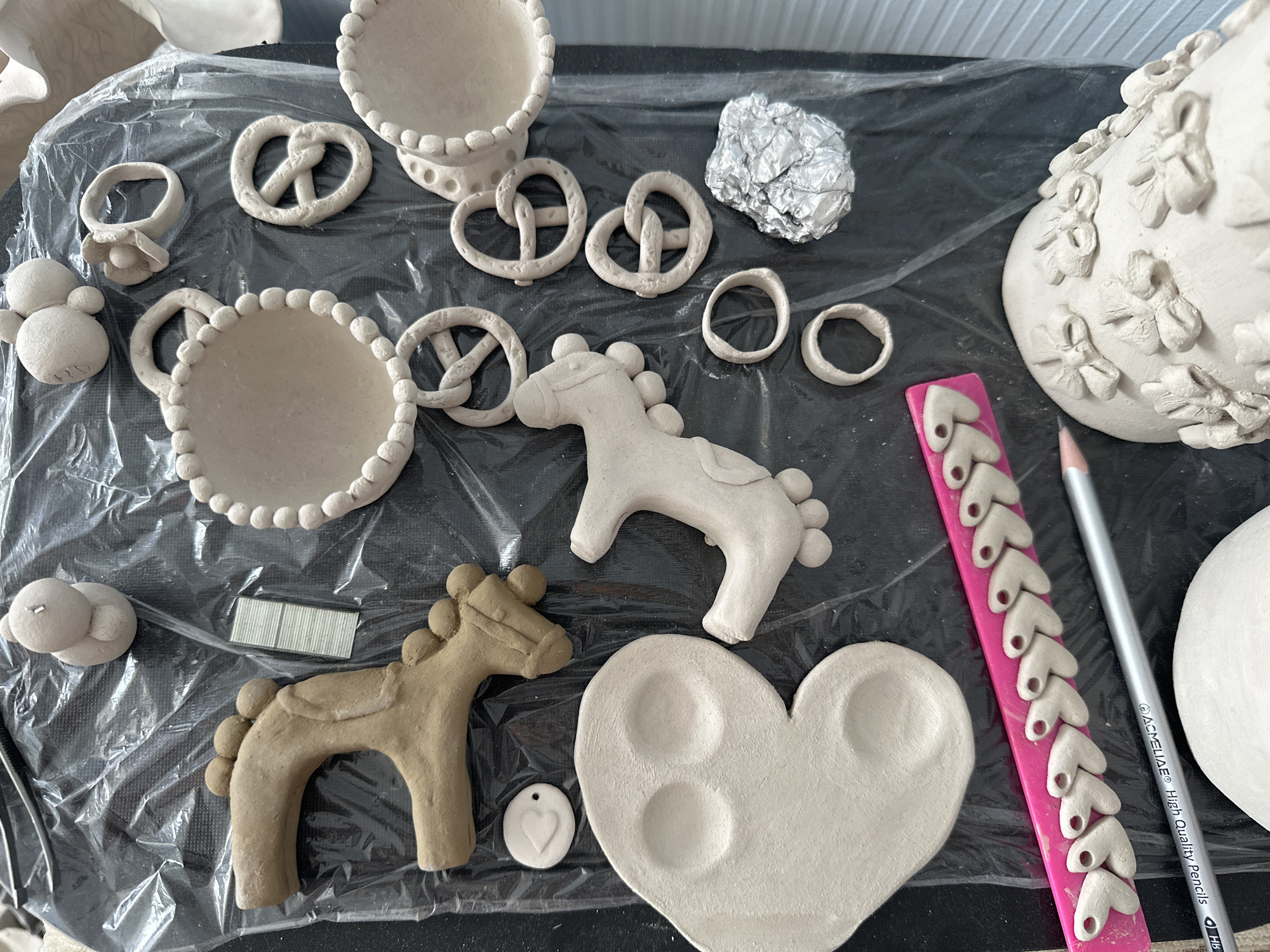 The image shows a collection of unfired clay objects laid out on a black plastic surface. These objects include several pretzel-shaped figures, two round dishes with textured rims, a heart-shaped object with two indentations, a stylized foot or leg structure, a small fish shape, and a bird-like figure made of stacked balls. There are also several ring shapes, a crumpled piece of aluminum foil, a pink object with a row of small heart shapes, and a pencil. Additionally, a larger, undecorated clay object with bow-like embellishments is partially visible. The lighting suggests an indoor setting. This image is not geotagged and therefore cannot be definitively located in Nevinnomyssk, Russia.