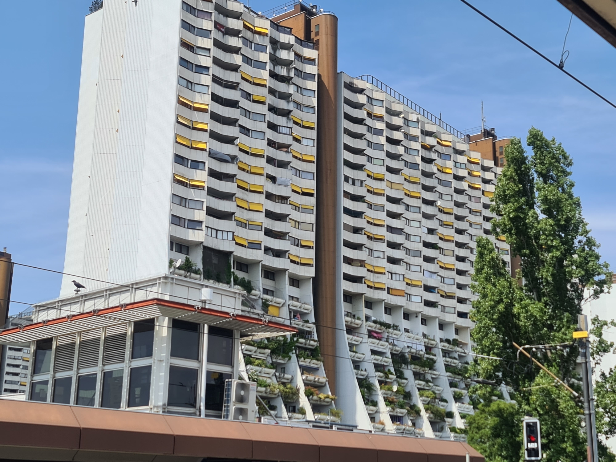 The image depicts a large, multi-story apartment building in Vienna, Austria, characterized by its distinctive curved balconies adorned with numerous plants and yellow awnings. The building's facade is white and grey, with a prominent, tall, rounded brown column running vertically through its center. The architecture suggests a Brutalist or similar modernist style. In the foreground, a section of what appears to be a tram or elevated structure is visible, with a red-brown colored roof-like overhang and large glass windows. On top of this structure, a bird is perched on a railing. Adjacent to the apartment building, to the right, are lush green trees. The sky is clear and blue, indicating a sunny day. The scene is urban, with hints of greenery provided by the trees and balcony plants, juxtaposed against the imposing concrete structure of the building. A traffic light showing red is visible in the lower right corner, along with overhead wires suggesting public transportation or power lines.