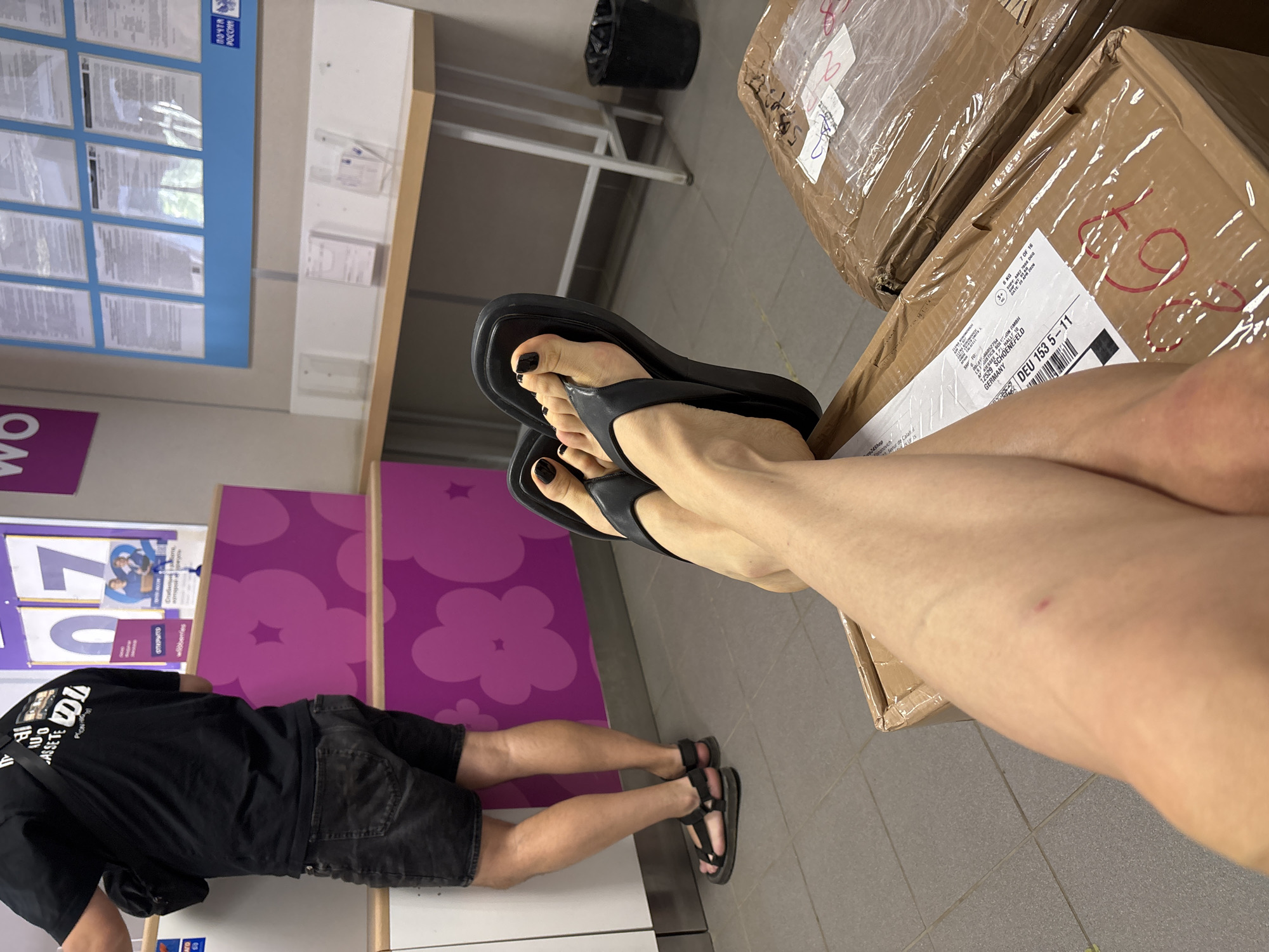 Here is a description of the image:
The image shows a person with legs crossed, with one leg resting on a brown cardboard box. The person is wearing black flip-flops on one foot and black sandals on the other. The person has black nail polish on their toes. The person's other leg is wearing a black sandal. The person is wearing black denim shorts and a black t-shirt. The background includes a purple wall with cloud and star designs, a white counter, and a blue and white poster. There are also other cardboard boxes visible. Writing on the box reads "GISTICS SOLON GMBH", "12529 SCHOENEFELD", "Sepur de Cala", "GERMANY", "DEU 153 5-11", and "6 κα". The word "wildberries" appears on the wall. A poster on the wall says "ОТКРЫТО".