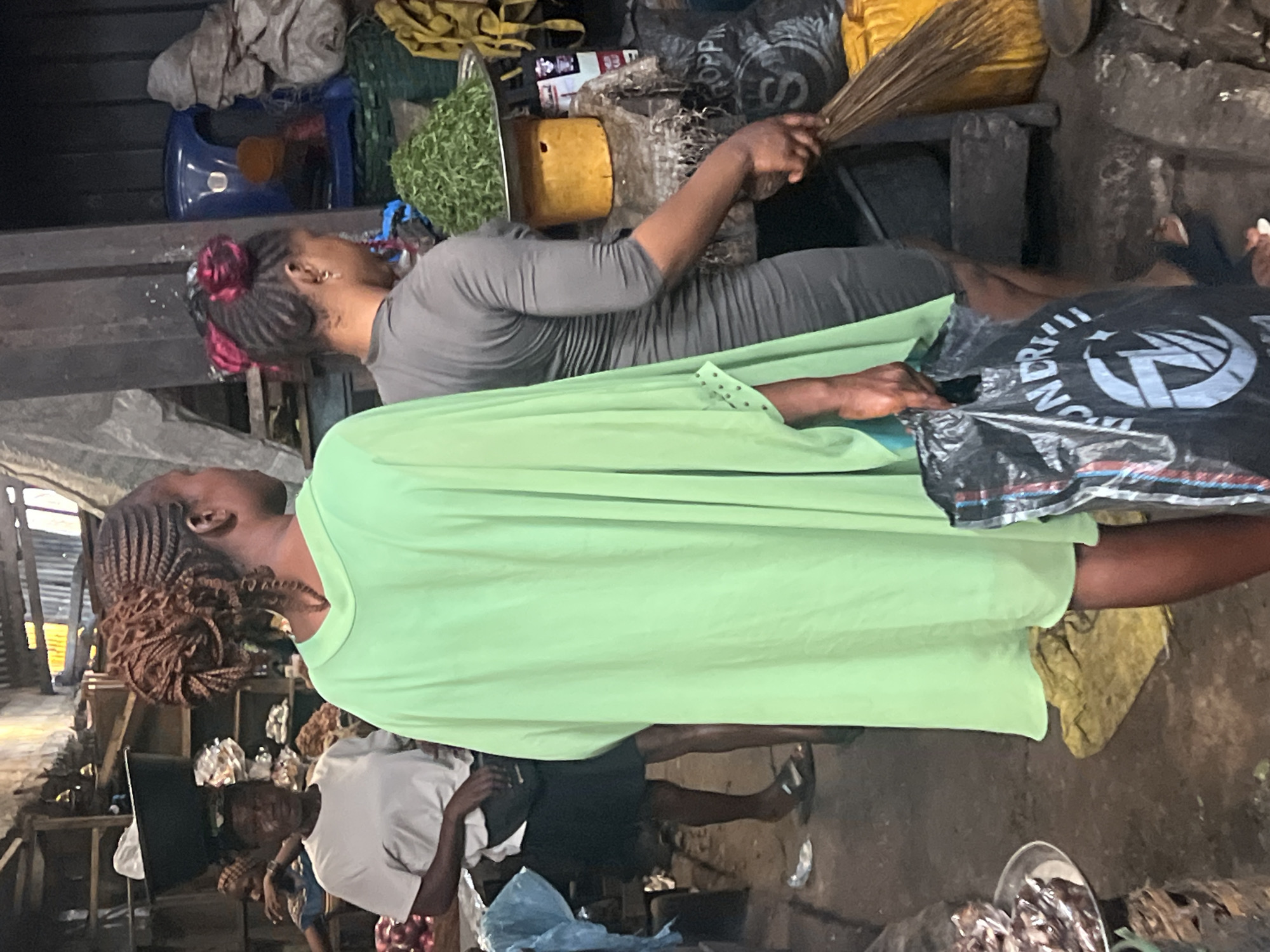 Two women are the primary subjects. One wears a gray dress and has a broom in her hand near various containers of food. The other is wearing a bright green dress. This scene appears to be in a market environment in Nkpor, Nigeria, with other individuals and market stalls visible in the background.
