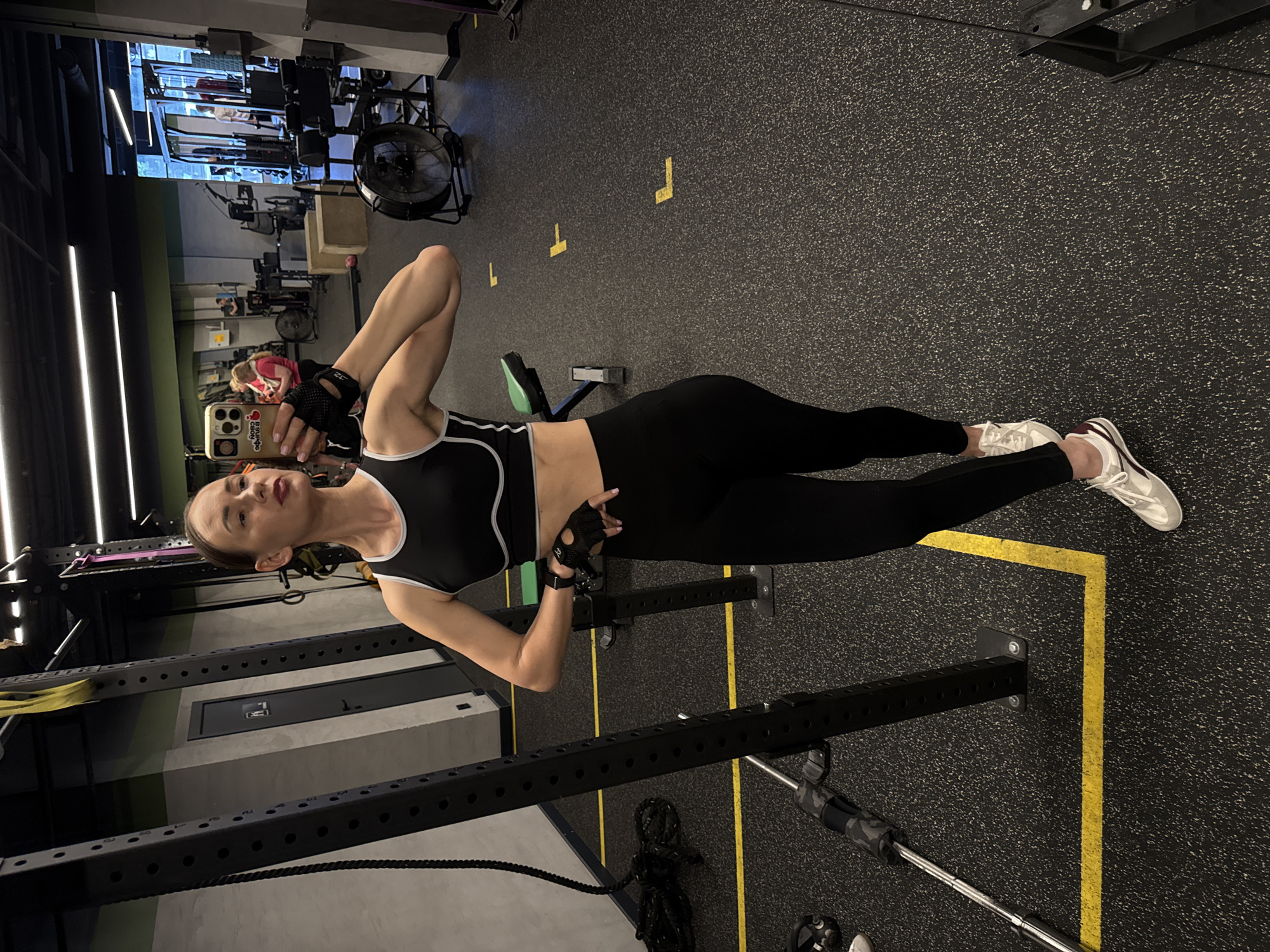 A woman poses for a selfie in a modern indoor gym, located in Sochi, Russia. She is standing upright, looking towards the camera, holding an iPhone in her left hand. The phone features a cream-colored case with a red cherry design and Cyrillic text, including the word "Яблоко" (Apple).
The woman is dressed in black athletic wear, consisting of a black sports bra with white trim and black form-fitting leggings. She wears white sneakers with maroon accents and black fingerless workout gloves. Her hair is styled back, and she has a calm, confident expression.
The gym floor is made of a dark, speckled rubber material with bright yellow L-shaped and straight line markings. Surrounding the woman is various gym equipment, including robust black metal structures resembling a squat rack or power cage, a battle rope coiled on the floor, and a barbell. In the background, other exercise machines, such as a cable machine, and a large industrial fan are visible. The ceiling is equipped with modern, linear fluorescent strip lights, providing bright illumination to the facility. Another person is partially visible in the distance among the equipment. The setting conveys a well-maintained and functional fitness environment.