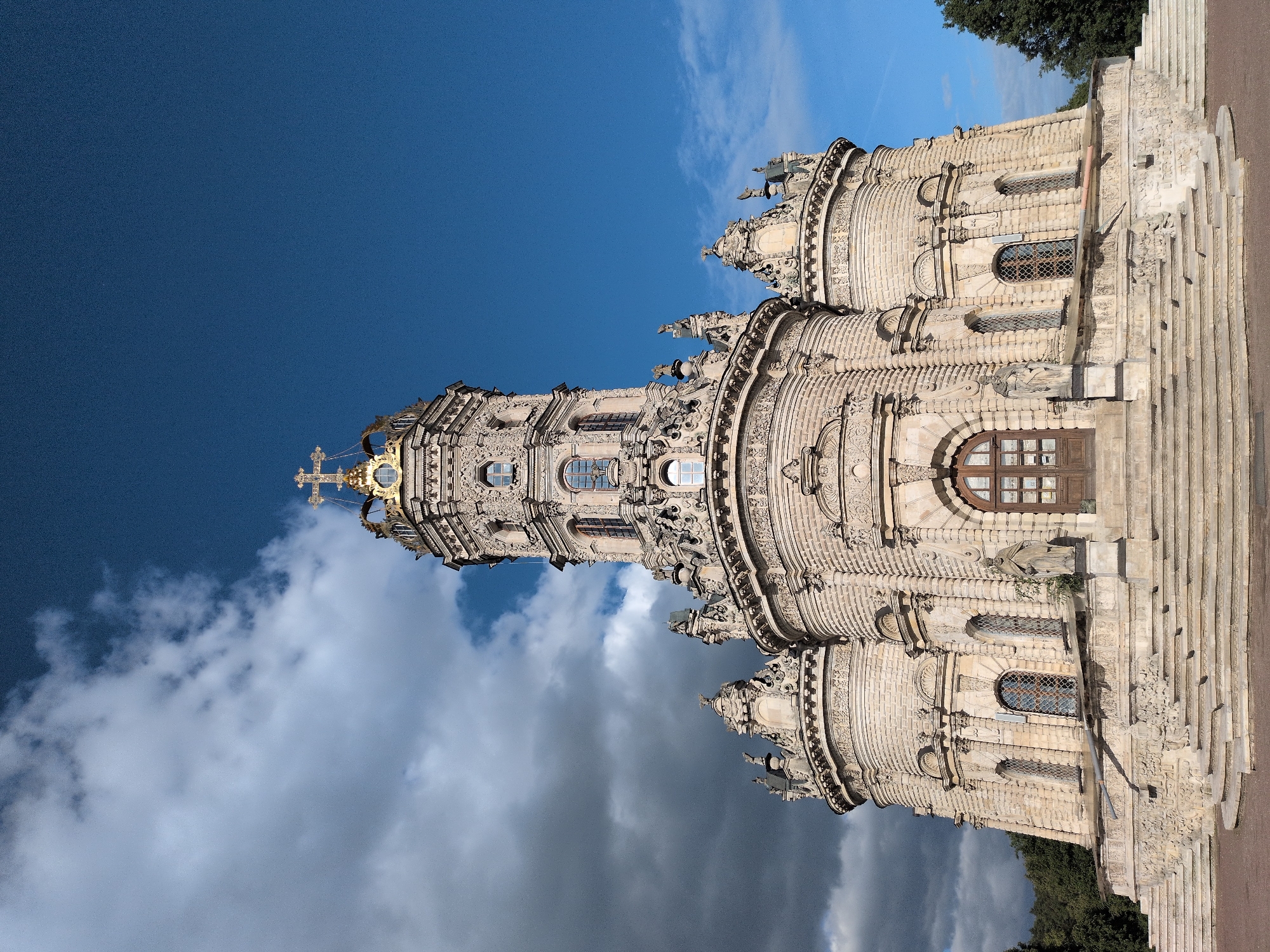 The image showcases the Church of the Sign of the Holy Virgin in Dubrovitsy, Russia. The church is an ornate baroque structure, with a towering central spire topped by a golden cross. Intricate carvings adorn the exterior of the church, featuring a series of arches, windows, and decorative elements. A large wooden door is visible on one of the church's rounded sides. The church is constructed of light-colored stone or brick. The sky behind the church is divided between deep blue above and a layer of billowing clouds below. The ground near the base of the church is made of paving stones. Green foliage appears at the edges of the image.