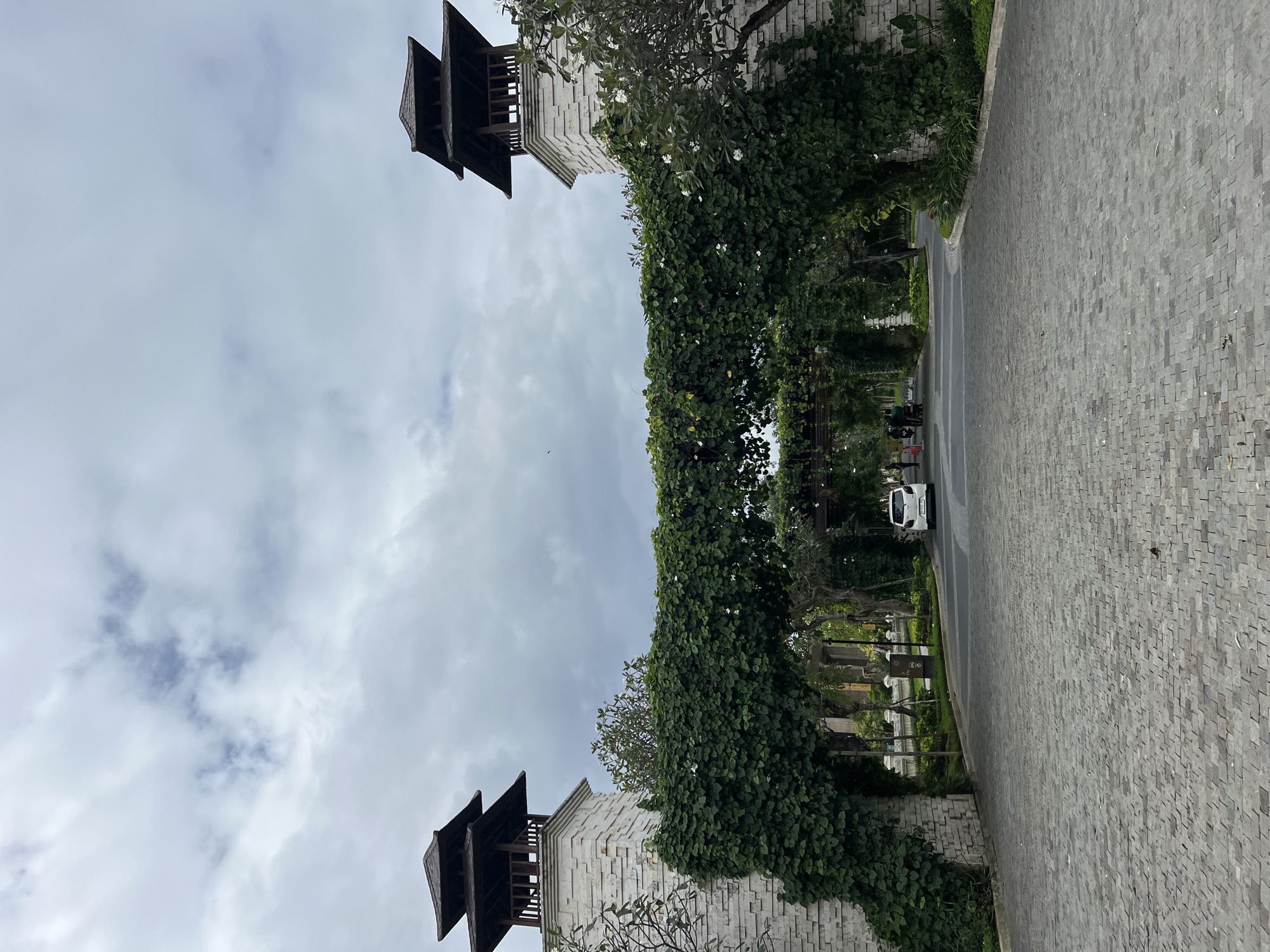 The image shows a paved entranceway to what appears to be a resort or complex in Denpasar, Indonesia. The entrance is flanked by two stone towers topped with tiered, dark wooden roofs. A thick, green vine covered in small white flowers arches over the roadway, connecting the two towers. The sky is overcast with scattered clouds. A white car is visible on the road in the distance, and a few people and a motorcycle are also present further down the path. Lush greenery and trees line the sides of the entrance.