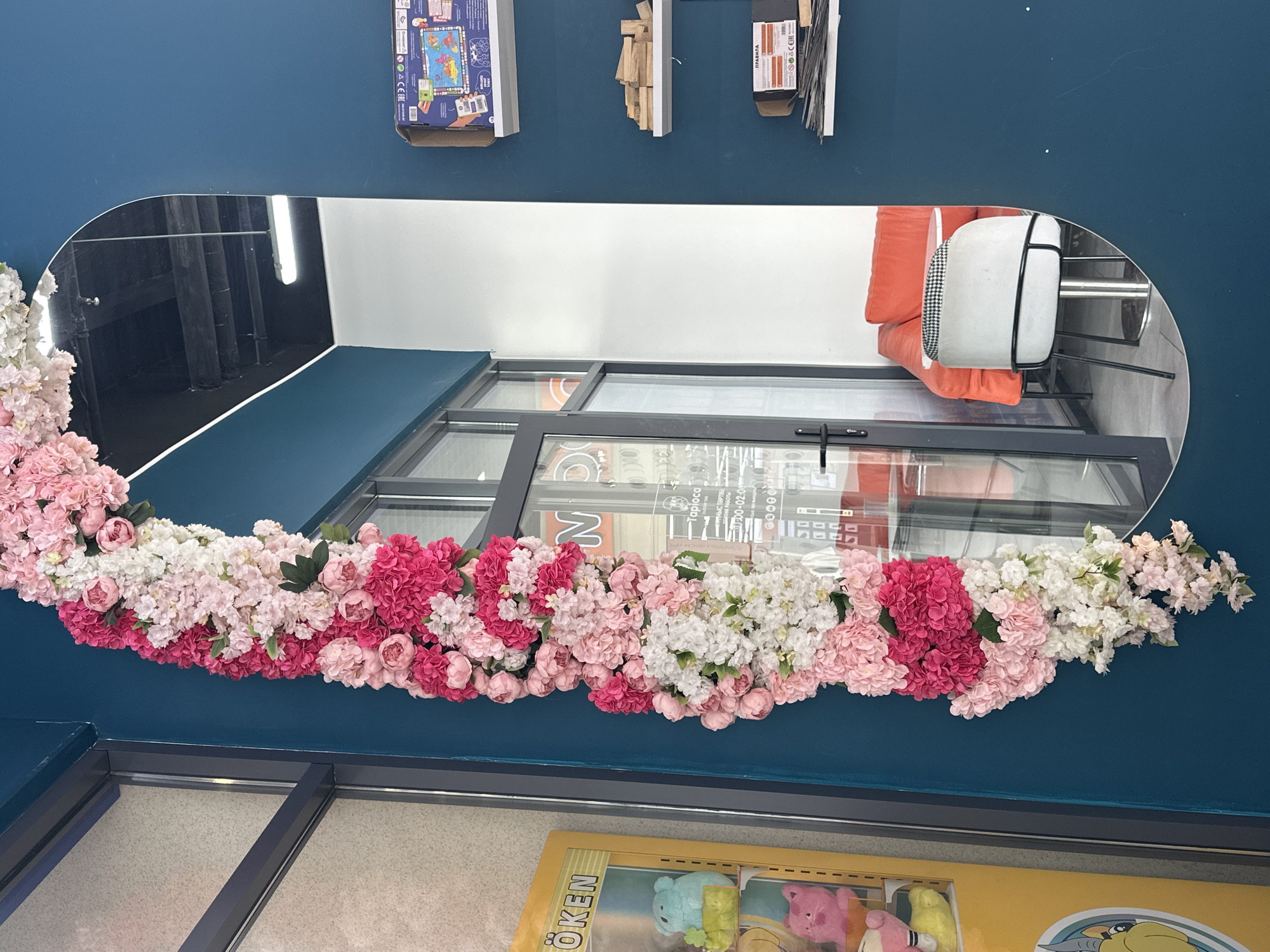 A tall, oval mirror on a teal wall is decorated with a cascade of artificial flowers in shades of pink, white, and fuchsia. To the left of the mirror, a yellow gumball machine with stuffed animals is visible behind a window. The mirror reflects a room with a bright orange sofa, a white chair with a black and white patterned seat, and a small round table. Through a glass door in the reflection, text on a sign appears to be in Cyrillic script. The overall scene suggests an interior space, possibly a shop or cafe, in the city of Aktau, Kazakhstan.