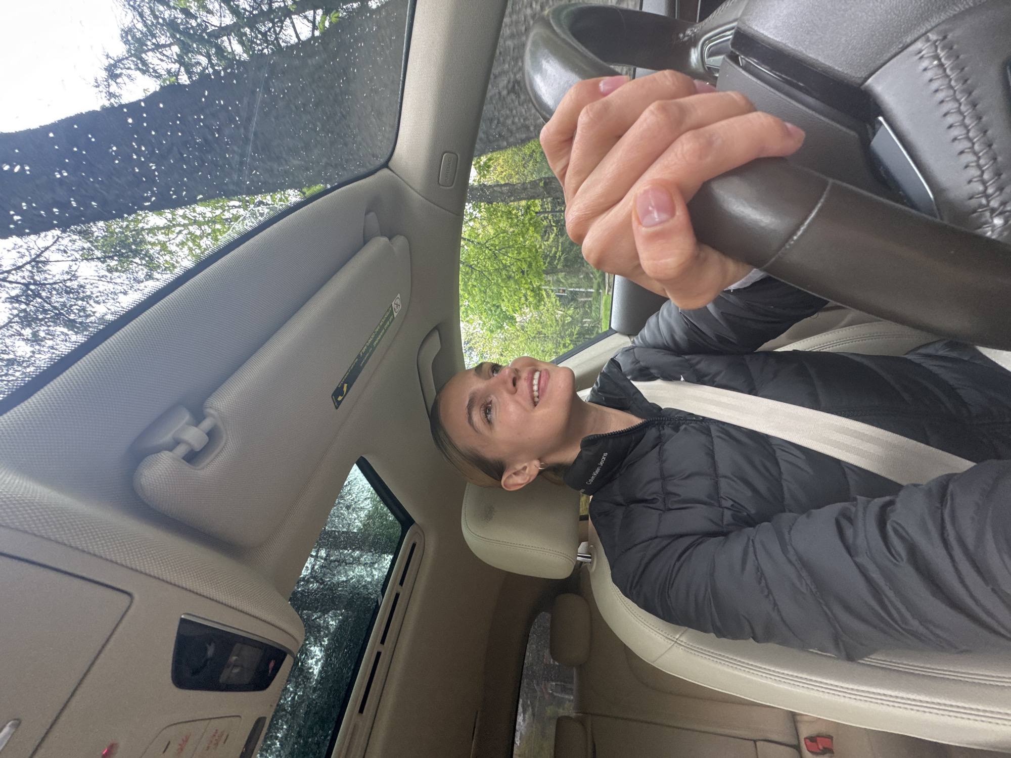 An indoor shot taken from inside a car captures a young woman in the driver's seat, looking upwards with a happy, smiling expression. She has light brown hair, fair skin, and natural-colored nails, and is wearing a black puffer jacket with "Calvin Klein Jeans" visible on the shoulder. A beige seatbelt is fastened across her chest. Her right hand rests on the dark, textured steering wheel.
The car's interior features a light beige or cream color scheme for the headliner, sun visors, and leather seats. A visible "AIRBAG" warning label is present on the passenger-side sun visor, along with other smaller text. An overhead console with various buttons and indicator lights is also visible.
Through the car's windows and sunroof, the outdoor environment is visible, dominated by the green foliage of trees, suggesting a natural or wooded setting. Numerous water droplets are present on the glass surfaces, particularly the windshield and sunroof, indicating recent rain or an ongoing drizzle. The scene is well-lit by natural daylight. The general atmosphere conveys a sense of ease or happiness during a drive or stop in Tbilisi, Georgia.