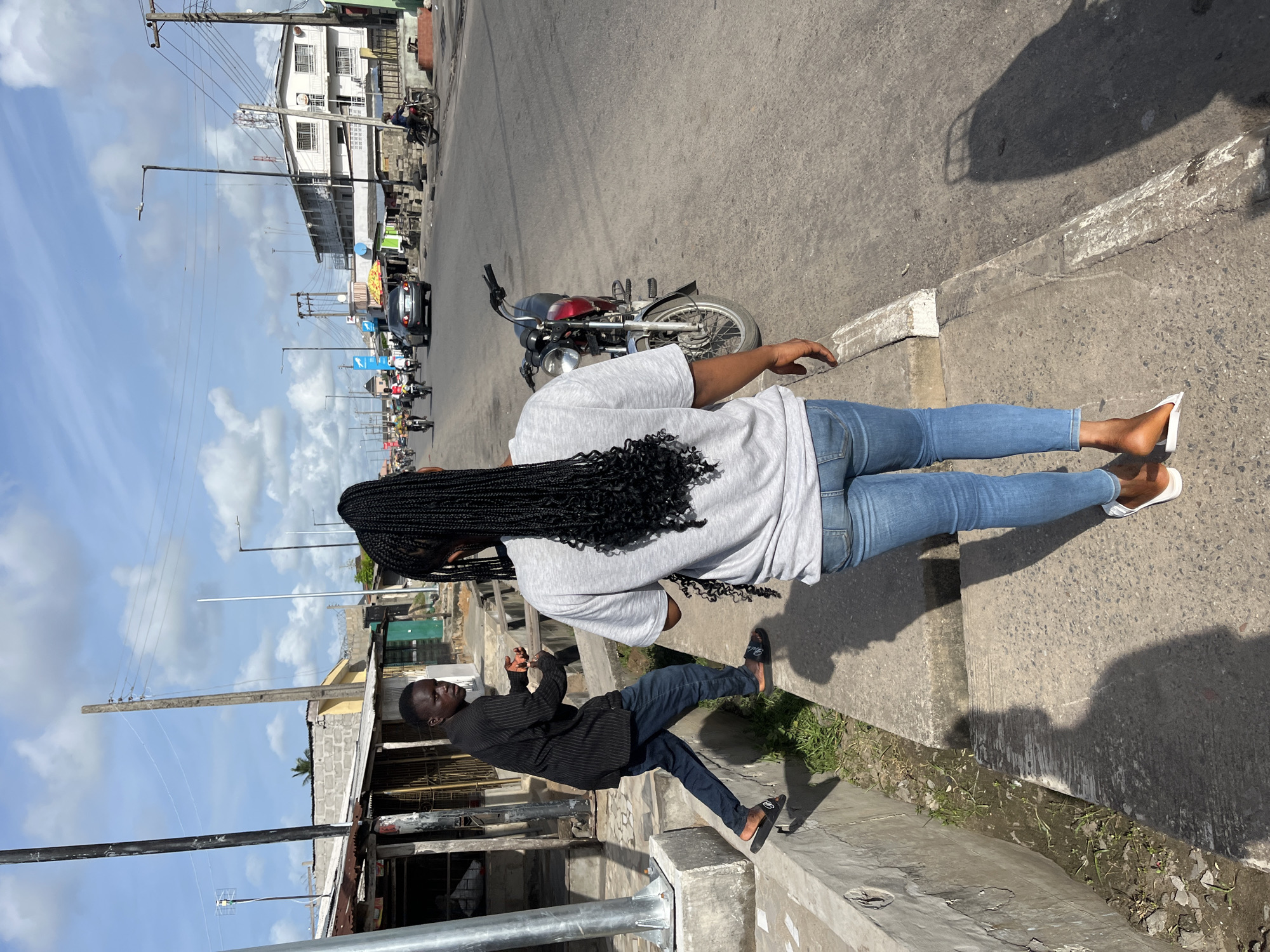A street scene in Badagry, Nigeria, shows two individuals on opposite sides of a raised curb. A female with long, dark braids wears a gray shirt, blue jeans, and white sandals while standing on the road sidewalk with a motorcycle parked near her. A male in a dark striped shirt and dark pants stands on the building sidewalk. In the background, utility poles, buildings, traffic, and a blue sky with scattered clouds are visible.