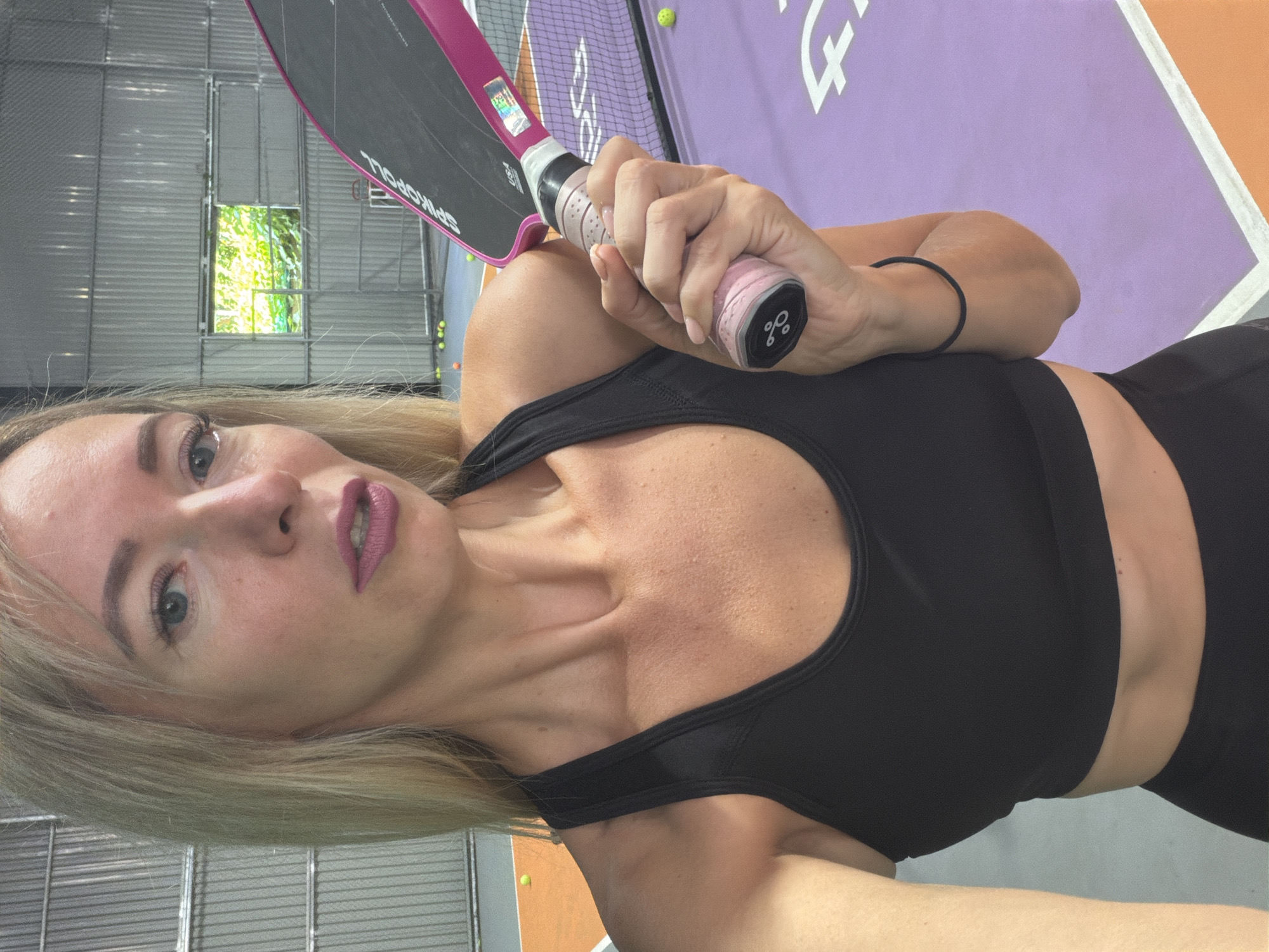 A woman with blonde hair and light-colored skin is holding a pink and black paddle. She is wearing a black sports bra and black athletic bottoms. The background appears to be an indoor sports facility with metal walls and a window showing greenery. The floor is colored in orange and purple sections, and there are tennis balls visible on the court. The woman is looking towards the camera with a neutral expression, and her left hand is visible holding the paddle. The scene is well-lit, and the focus is on the woman and the paddle. There are no other people visible in the image. The specific location in Da Lat, Vietnam, cannot be determined from the provided image alone.