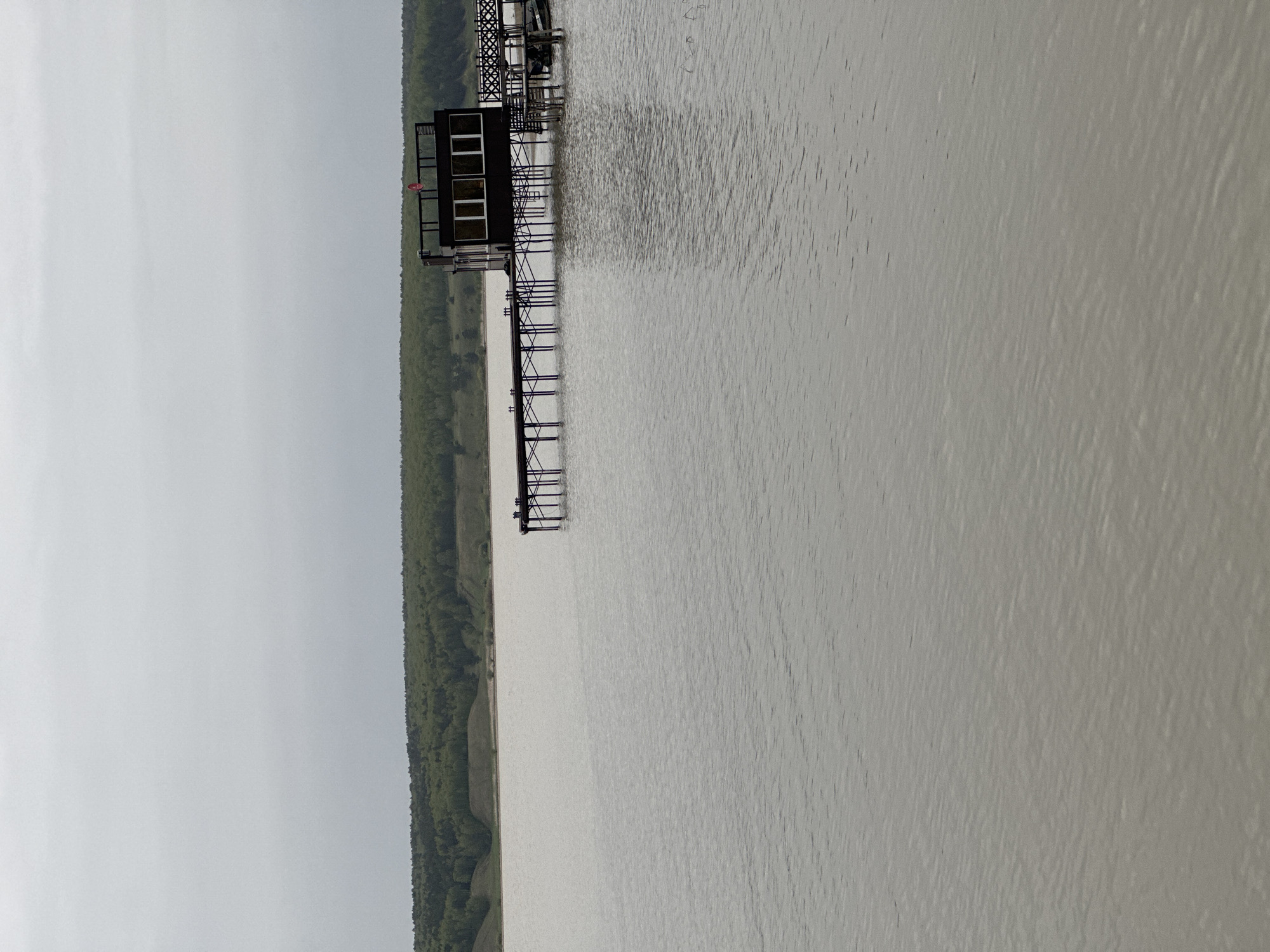 The image shows a structure on a body of water, possibly a river or lake, with a forested shoreline in the background. The structure is supported by metal stilts extending into the water. The weather appears overcast. Based on the request for the location in Aviator, Russia, the structure is likely a fishing pier or small platform located on the Volga River or one of its tributaries in that region. No people are visible.