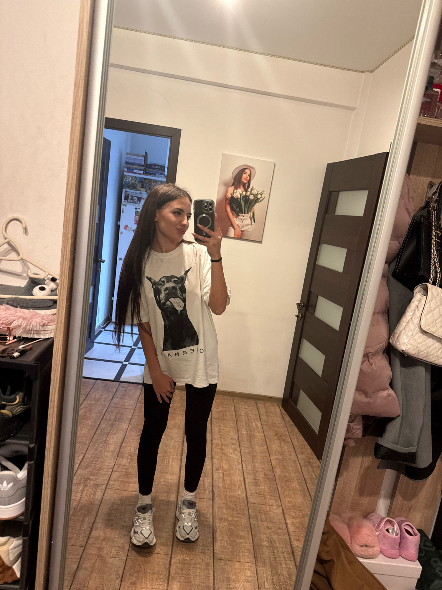 A young woman with long, dark hair takes a mirror selfie in a hallway. She is wearing a white t-shirt with a large black dog graphic and text that reads "КАМЯЗО". She's also wearing black leggings and white athletic shoes with grey accents. The woman is standing in front of a full-length mirror, reflecting a room with wooden floors. To her left, a tall, narrow shelf unit is partially visible, filled with shoes and other items. To her right, a dark wooden door with frosted glass panels is visible, as is a closet with hanging clothes and bags, including a white quilted handbag. On the wall above the door, a framed picture of a woman holding a bouquet of white tulips is displayed. The lighting suggests it is daytime indoors. The setting appears to be a home in Chișinău, Moldova.