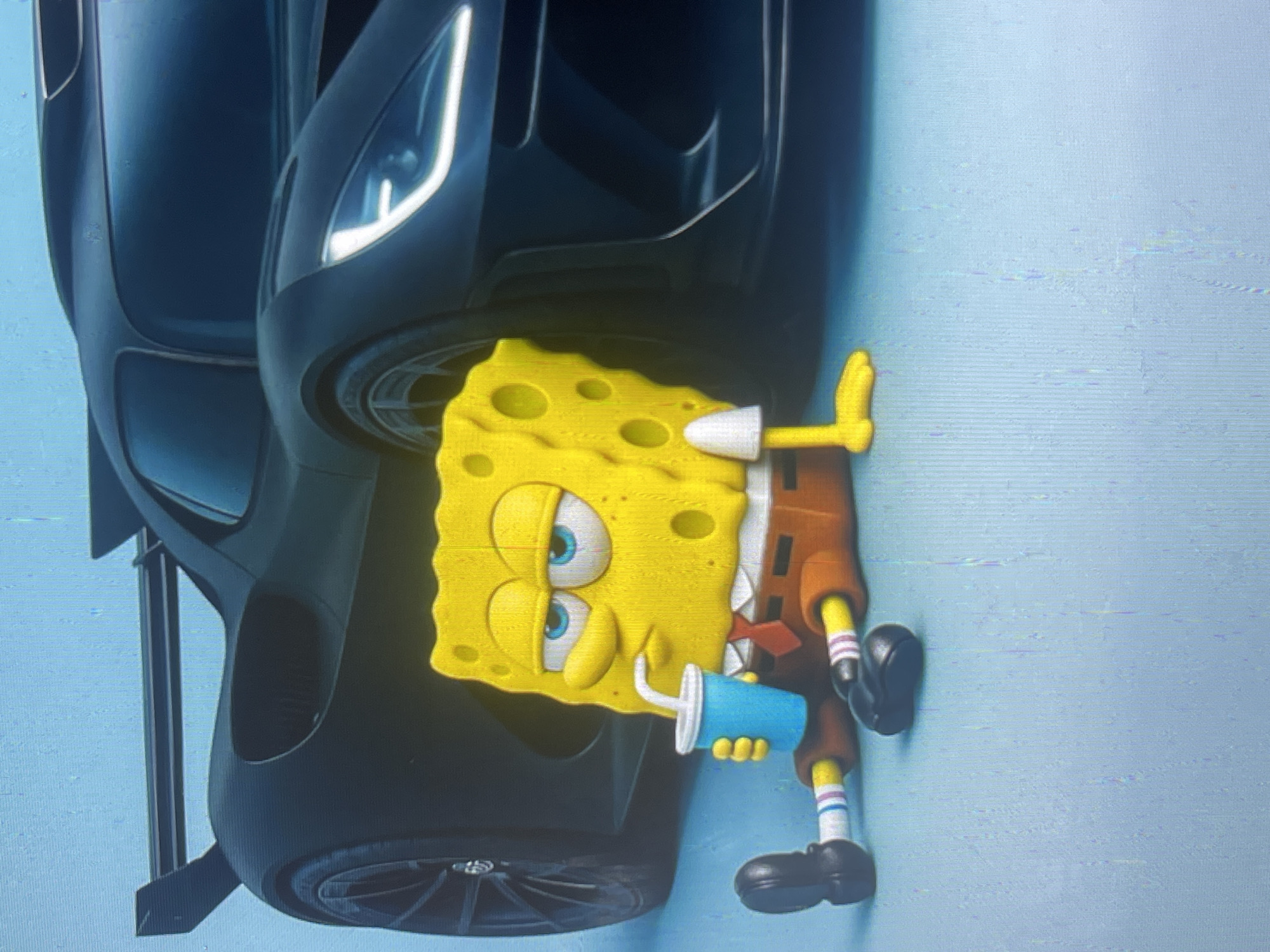 A still image shows a cartoon character, SpongeBob SquarePants, sitting on a light blue surface. He is leaning against a dark-colored sports car. SpongeBob is wearing his usual square pants, shirt, and tie, and holding a blue cup with a straw. His expression is relaxed, with his eyes half-closed. The sports car appears to be modern and sleek, with a large spoiler and distinctive headlights. The background is a plain, light blue color. There are no indications of the city of Bishkek City, Kyrgyzstan, in the image.