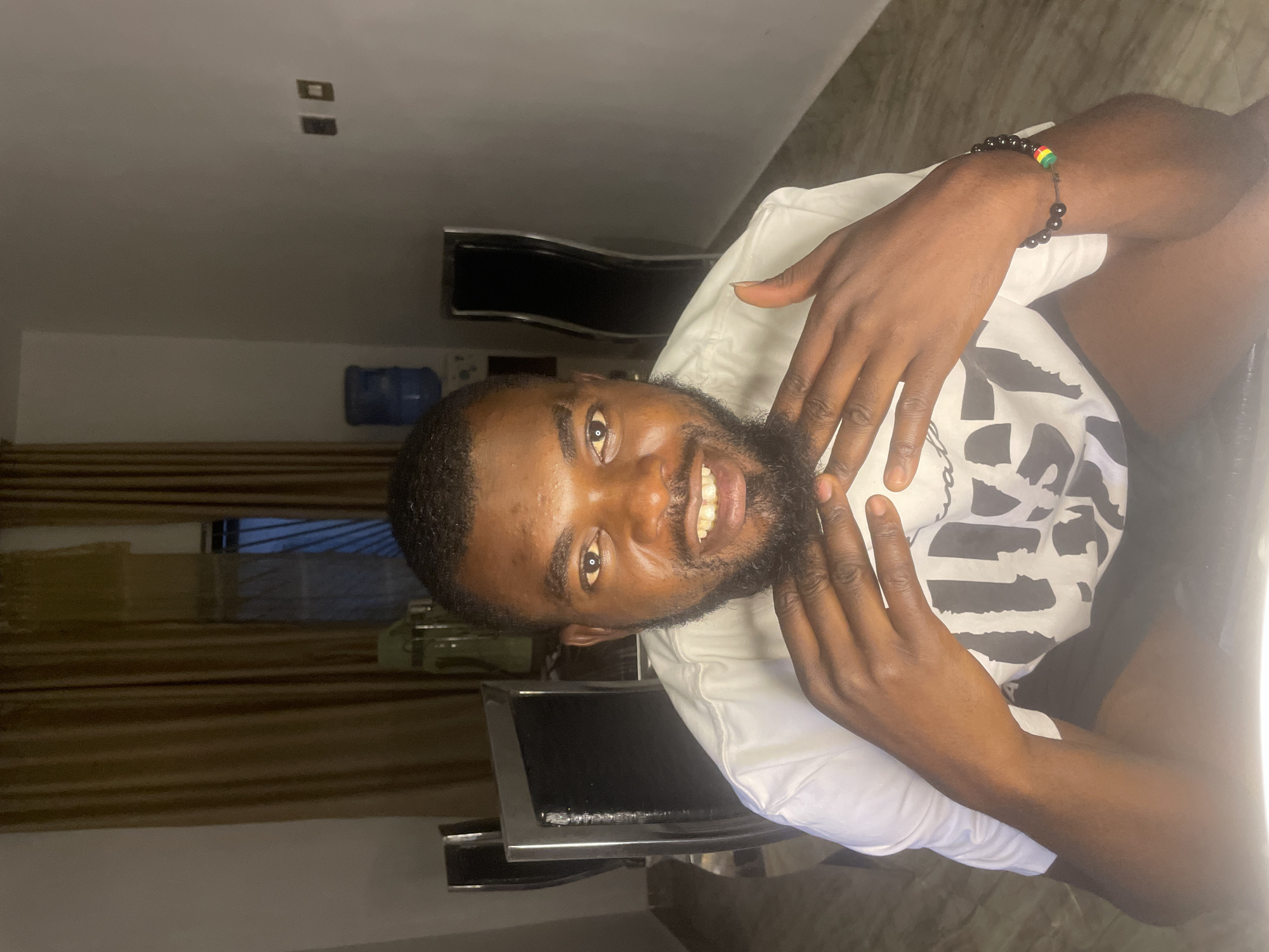 A Black man with dark hair and a beard is smiling directly at the camera. He is wearing a white t-shirt and a black bracelet with a green, yellow, and red bead on his left wrist. His hands are raised and positioned near his face. He appears to be indoors in a room with neutral-colored walls. A window with blinds can be seen in the background. A black chair is also visible. The image may be taken in Jos, Nigeria.