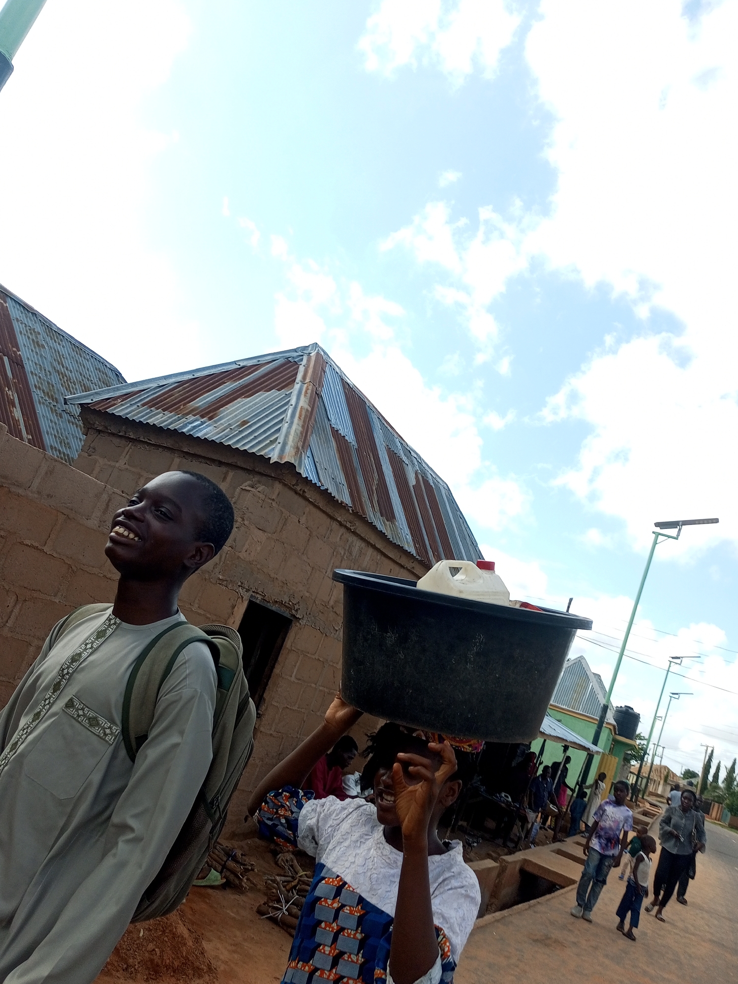 In Tempure, Nigeria, a young person is captured in a low-angle shot carrying a large black bucket filled with a white container on their head. The background features a street scene with people walking and buildings with corrugated metal roofs. The sky is bright blue with scattered white clouds, suggesting daylight. The person in the foreground, a young male wearing a light gray garment with intricate white patterns, is looking to the left and smiling. Another young person, also carrying a bucket on their head, is visible to the right, though their face is not clearly seen. The scene appears to be set in a bustling outdoor environment, possibly a market or a residential area, with a paved road and a drainage channel on the side.