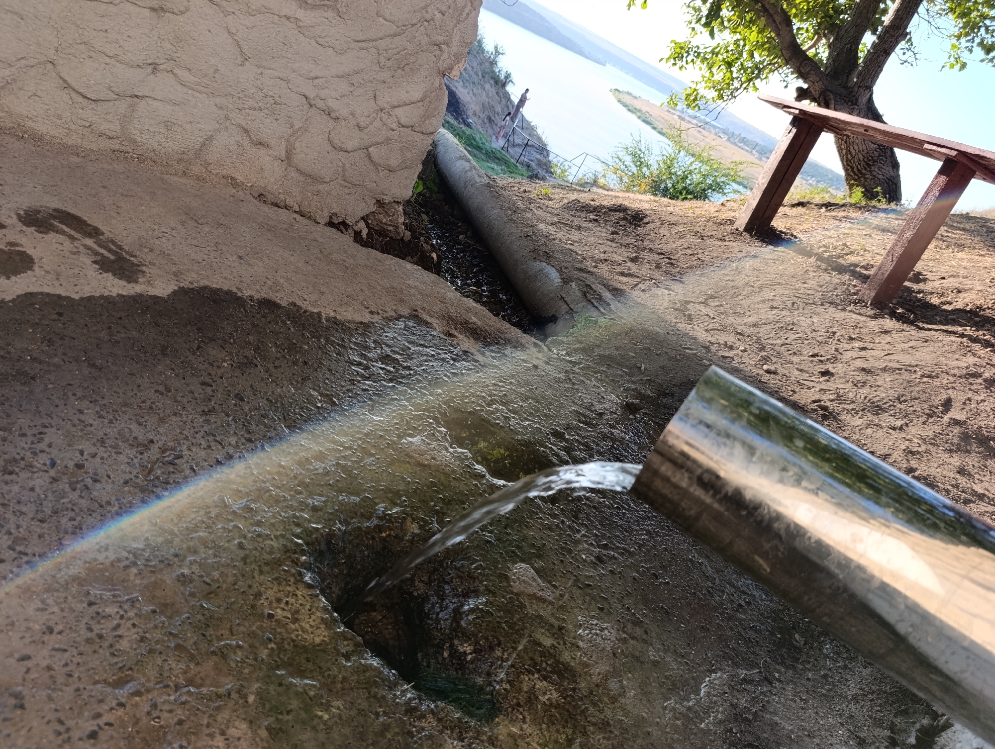 In Roghi, Moldova, a metal pipe is discharging water onto a concrete surface. A small rainbow is visible where the sunlight refracts through the water spray. The water flows into a dark opening in the concrete. In the background, a large body of water, likely a river or lake, is visible. A tree and a wooden bench are situated on the bank overlooking the water. The terrain is dry and dusty.