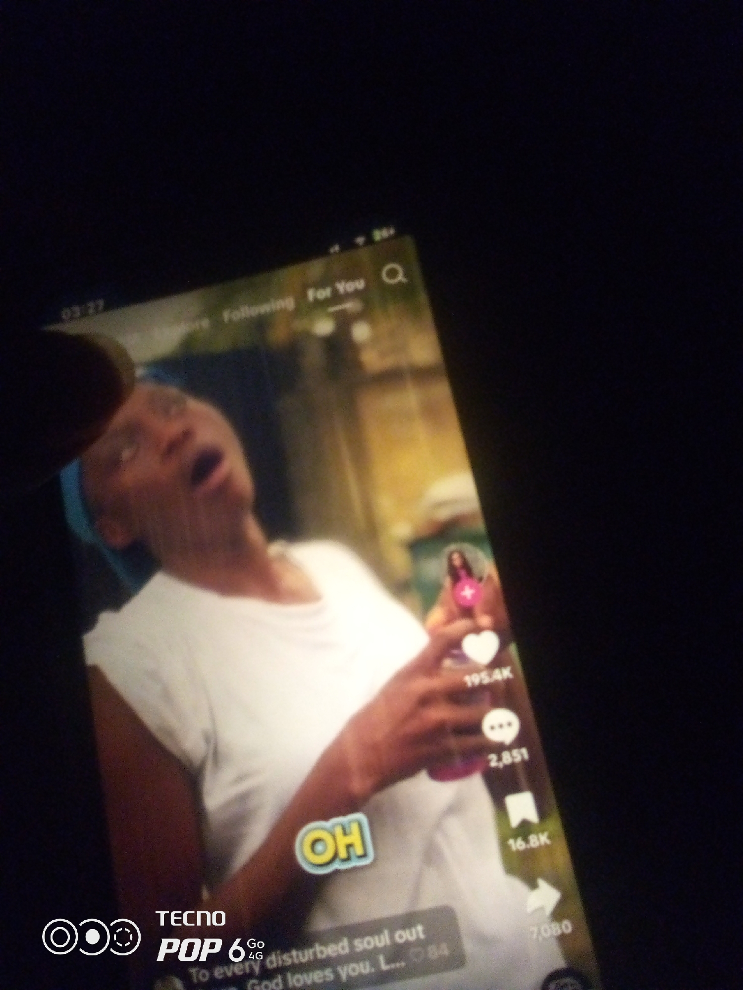 This is a close-up, dimly lit, high-angle shot of a smartphone screen displaying a video. A person's finger is visible in the upper left corner, obscuring part of the screen. The video appears to be a social media post, likely from a platform like TikTok, given the interface elements such as likes, comments, shares, and a "For You" indicator. The central subject of the video is a Black person, with their mouth open and eyes looking upwards, seemingly mid-expression. They are wearing a white t-shirt and a blue headscarf. Text overlays on the screen include "OH" in a stylized yellow font, and a caption that reads, "To every disturbed soul out. God loves you. L... 84". There are also statistics indicating 195.4K likes, 2,851 comments, and 16.8K shares. The Tecno logo and "POP 6 Go 4G" branding are visible in the lower left corner of the screen. The background of the video is out of focus and appears to be an outdoor setting with greenery. The image does not provide any specific information to determine the location within Howayi, Nigeria.