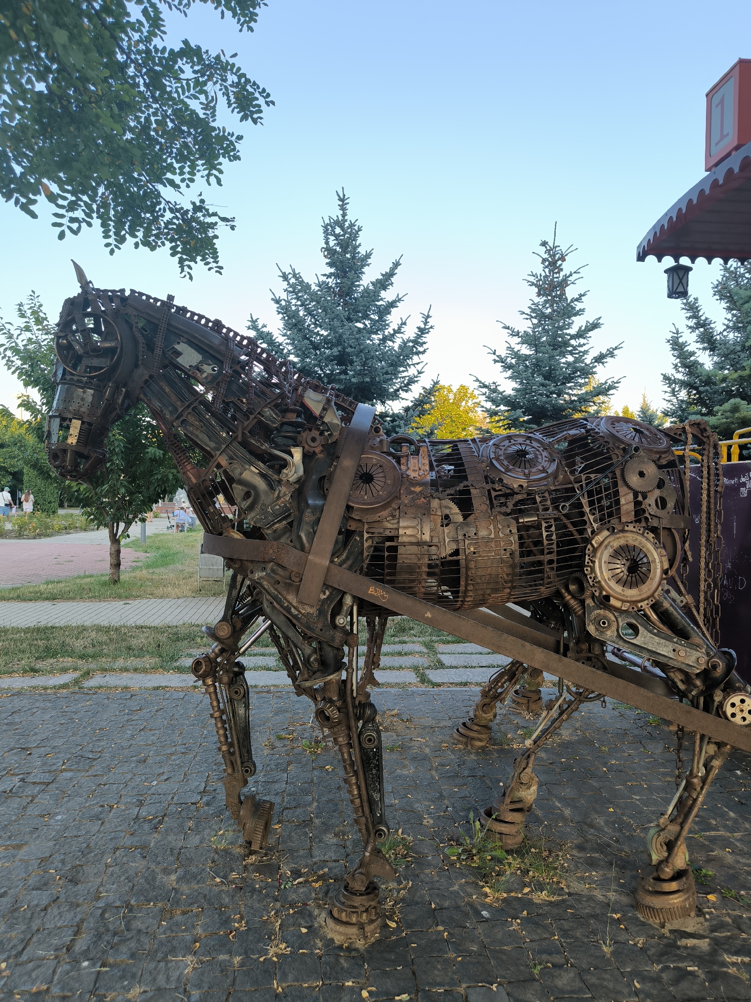 The image shows a large metal sculpture of a horse in a park-like setting. The sculpture is made from various discarded metal parts, including gears, chains, and engine components, giving it a rustic and industrial appearance. The horse sculpture is positioned on a paved walkway made of dark gray cobblestones. In the background, there are green trees, a clear blue sky, and a small structure with a red roof and a lamp. A few people are visible in the distance, walking in the park. The lighting suggests it is either early morning or late afternoon. The sculpture is likely a public art installation.
This artwork is located at the Sculpture Park in Chișinău, Moldova. Specifically, this sculpture is known as "The Horse" and is a notable piece within the park's collection of metal sculptures.