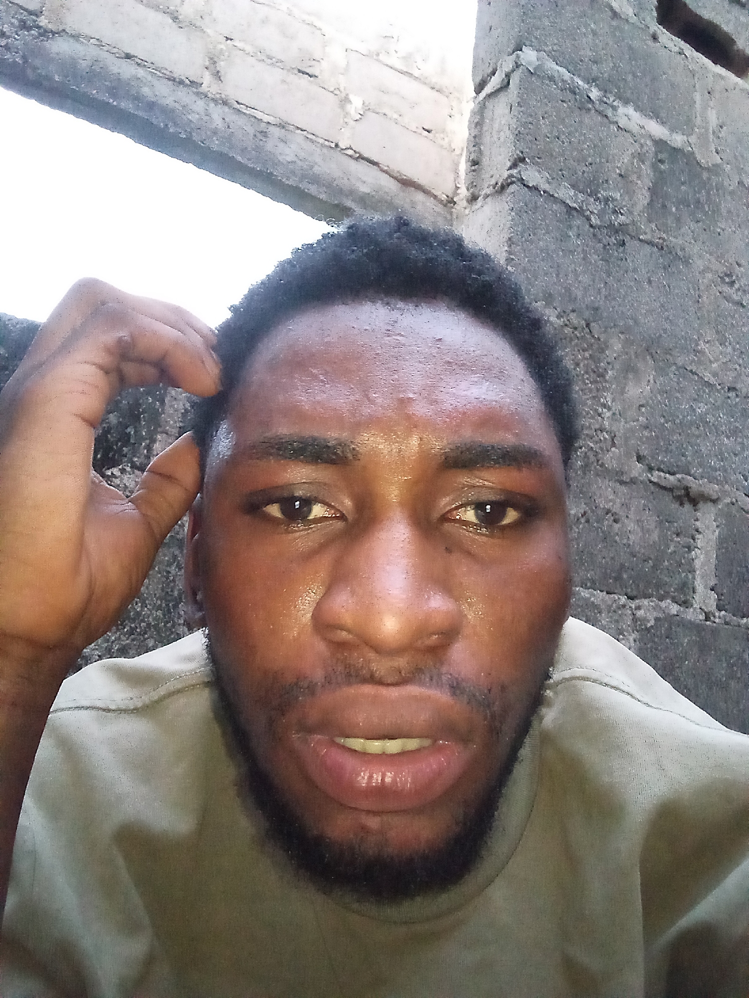 A selfie taken in Lokoja, Nigeria shows a man looking directly at the camera. The man is of African descent with dark skin and short, curly black hair. He is wearing a light brown t-shirt. His right hand is raised to his head, with his fingers touching his hair. His face is shiny, suggesting sweat. The background consists of a rough, grey brick wall and a bright, undefined area at the top left, possibly indicating an opening or sky.