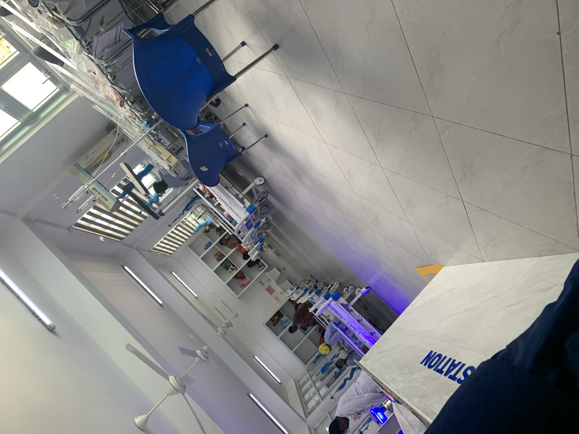 The image depicts an interior scene, appearing to be a hospital ward, in Dar es-Salaam, Tanzania.
The scene includes hospital beds, equipment, and medical personnel attending to patients. In the foreground is a table, with "STATION" written on it. The floors are tiled. The lighting appears bright. There are hospital chairs. Medical machines are present. Windows with sunlight present. Ceiling fans are observed. The view is angled.
