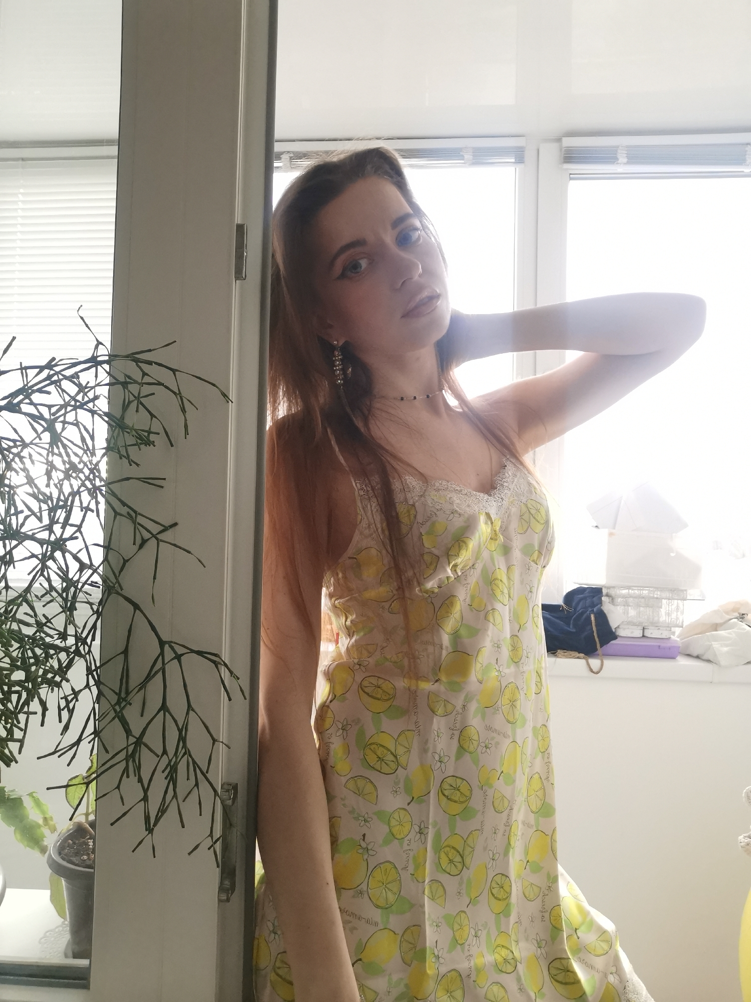 A young woman with long, reddish-brown hair is centered in the frame, posing indoors near a window or glass door. She looks directly at the viewer with a calm expression, her left hand resting behind her neck. She wears a light-colored slip dress or camisole adorned with a repeating yellow lemon and green leaf pattern, featuring white lace trim along the neckline. She has long, decorative earrings and a thin, dark choker necklace.
To the left, a large, intricate green houseplant with thin, branching stems (possibly a Rhipsalis) occupies the foreground. The background is brightly lit, indicating strong daylight coming through large windows fitted with horizontal blinds, which are partially visible on the left. The light creates a soft glow around the woman and brightens the overall scene, causing some areas in the background to be overexposed.
On the right side, behind the woman, a windowsill or ledge holds various items. These include stacked white objects (possibly boxes or folded fabric), a dark blue fabric item, a small purple container, and other less distinct articles.
The overall setting appears to be a residential indoor space, possibly a living room or bedroom with ample natural light. On the woman's dress, faint text is subtly integrated into the lemon pattern, appearing to say "natural" and possibly "lemon-calm" or similar words, though it is difficult to read clearly due to the fabric folds and pattern. The strong backlighting highlights the contours of the woman and the plant.