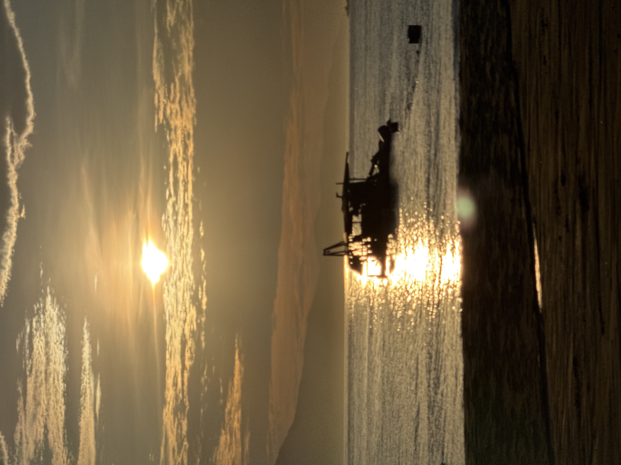 The image depicts a sunset over a coastal area in Đà Nẵng, Vietnam. A brilliant, low-lying sun is positioned in the upper-left quadrant, generating a strong lens flare and a prominent path of golden specular highlights across the calm water surface. The sky contains scattered clouds, illuminated by warm golden and muted grey tones. In the mid-ground, a heavily silhouetted utility boat, equipped with a superstructure and distinct rigging, is visible on the water. To the right of the boat, a dark, rectangular object floats. The distant background features a range of hazy, dark mountains spanning the horizon. The immediate foreground comprises a dark, sandy beach with a visible waterline where it meets the water. No human figures or discernible actions are present.