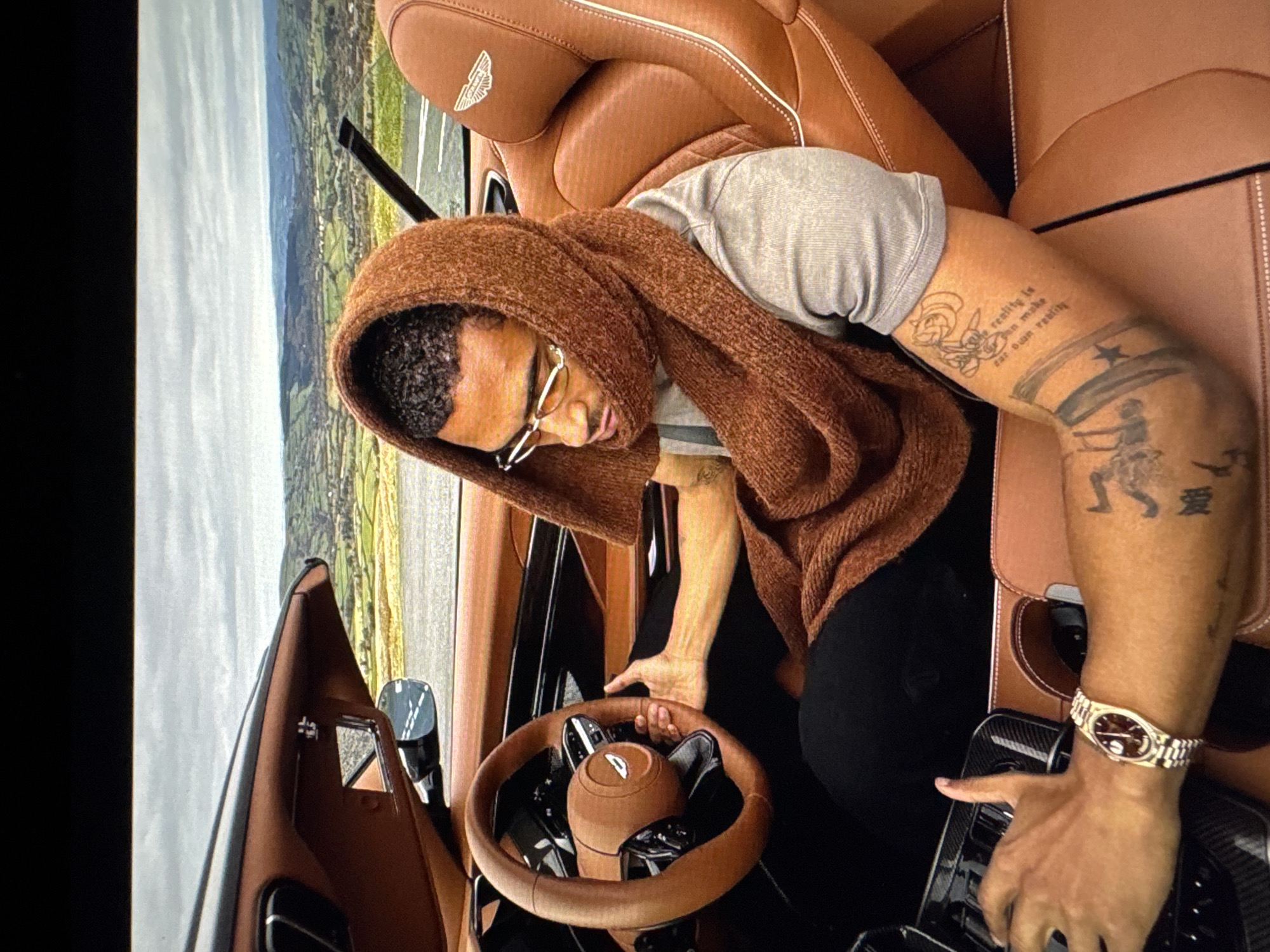 In a luxury convertible car with brown leather interior, a man wearing glasses and a brown scarf wrapped around his head is driving. The car's steering wheel is also brown leather. In the background, a landscape of rolling green hills and fields is visible under a cloudy sky. The man has tattoos on his arm, and he is wearing a gold watch. The scene does not provide any information about the city of Tallinn, Estonia.