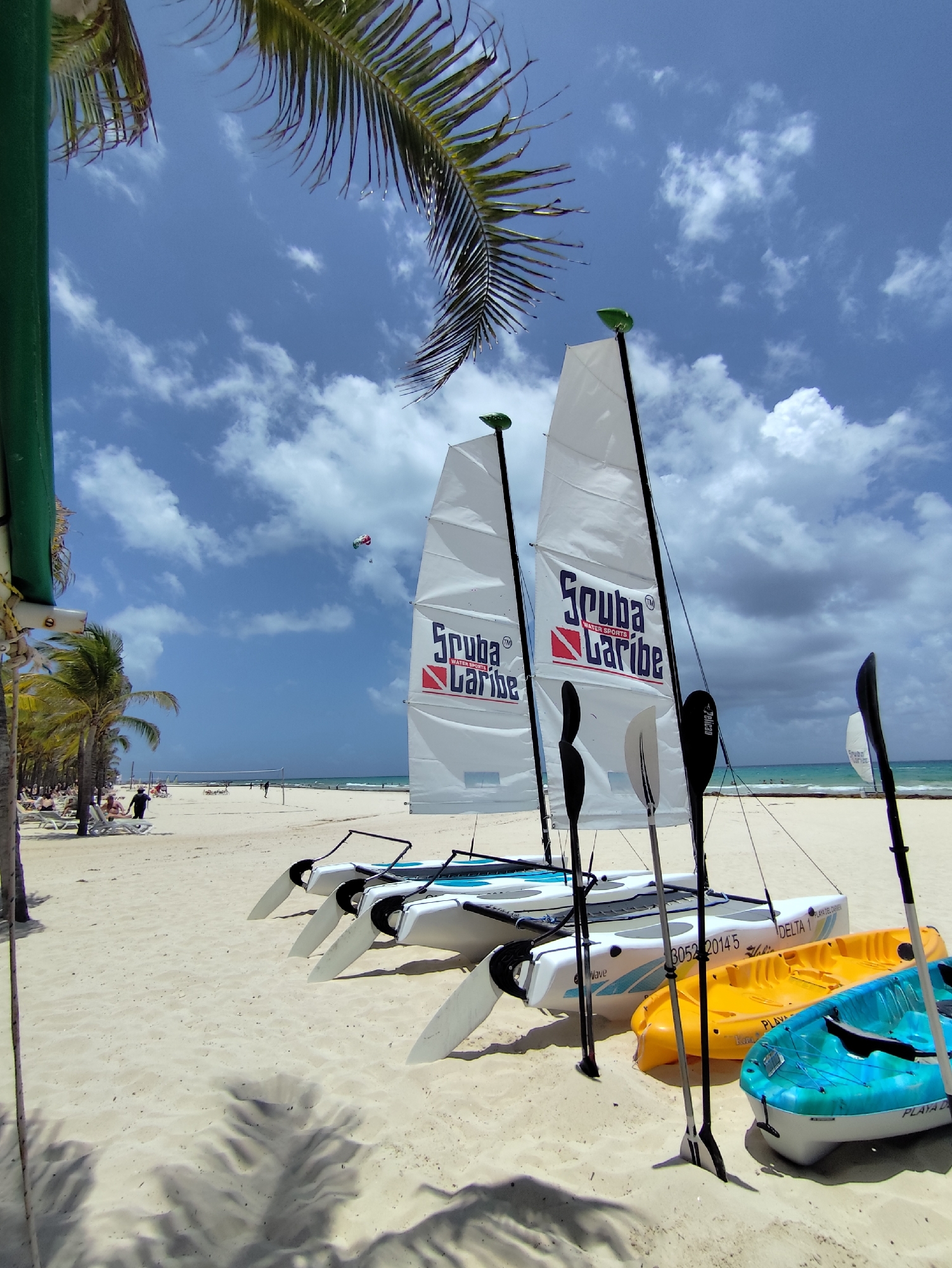 The image depicts a beach scene in Playa del Carmen, Mexico, under a sky with scattered clouds. In the foreground, there are two small catamarans with white sails bearing the logo "Scuba Caribe". Several paddles stand upright between the boats. To the right of the catamarans are three kayaks in yellow and turquoise colors. In the background, the beach is lined with palm trees and several people are relaxing on lounge chairs near the water. A person is walking along the shoreline. In the sky, a multicolored parachute is visible, suggesting parasailing activity. The sand appears light-colored and flat. The overall scene suggests a beach resort or water sports area.